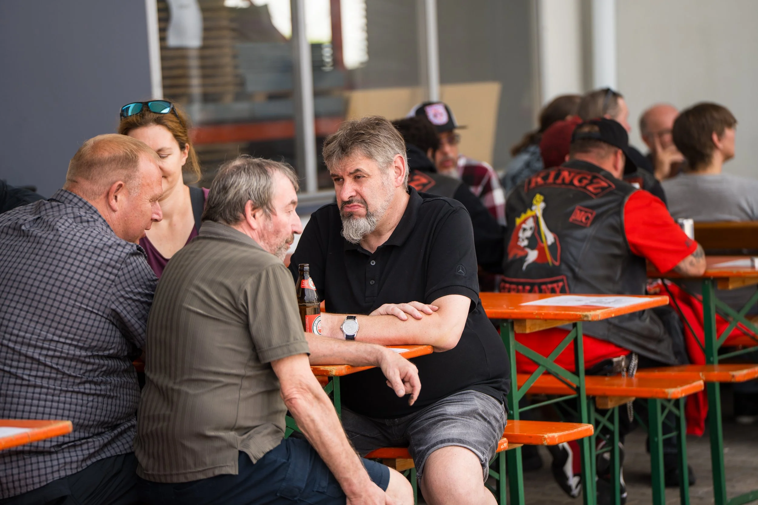 Menschen sitzen an einem langen Tisch im Freien, unterhalten sich und trinken Bier, während im Hintergrund weitere Personen am Tisch sitzen. Bei der Eröffnungsfeier von Gasolina Custom Motorcycles.