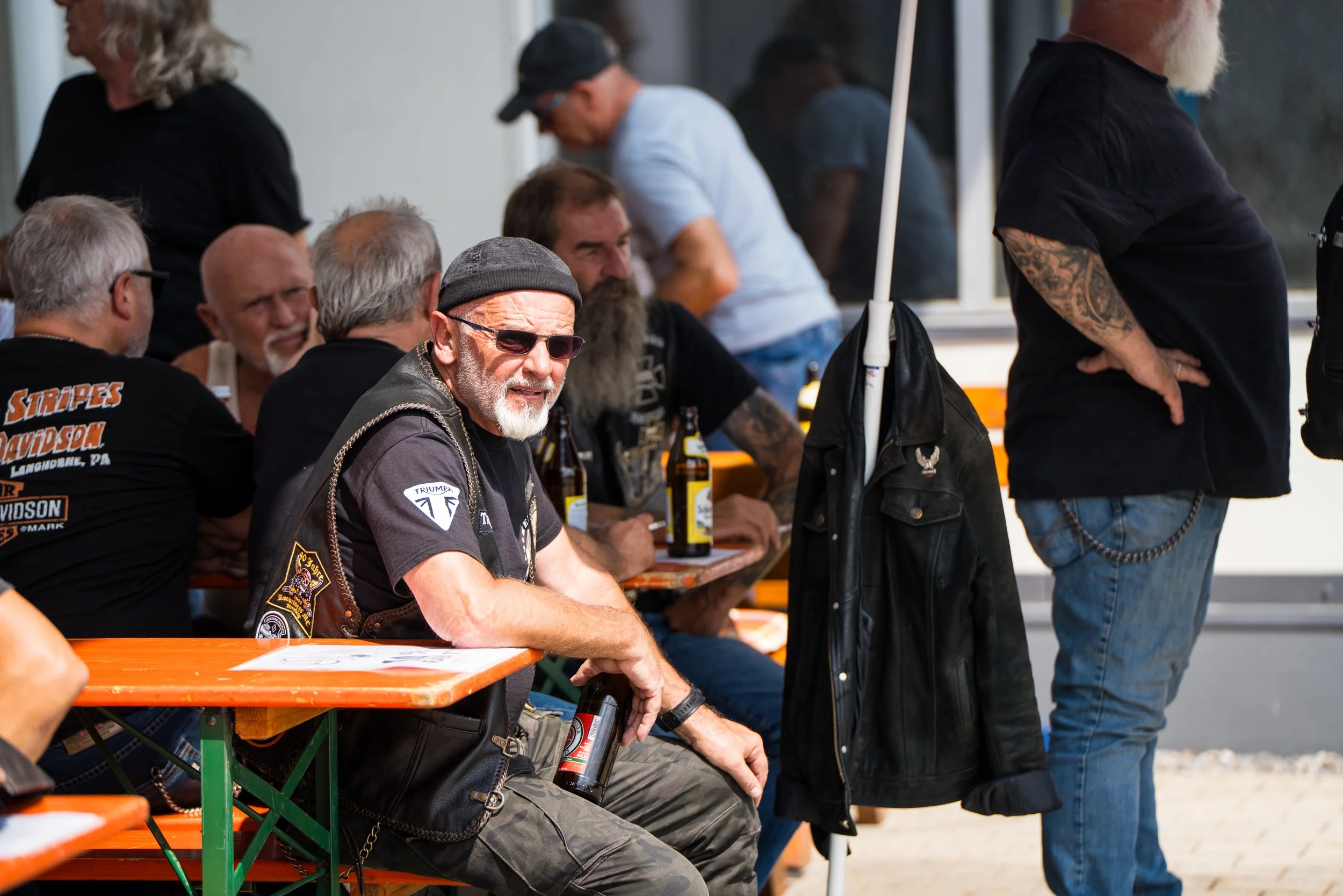 Mehrere Männer sitzen an einem langen Tisch im Freien, einige trinken Bier, während andere stehen.   Bei der Eröffnungsfeier von Gasolina Custom Motorcycles.