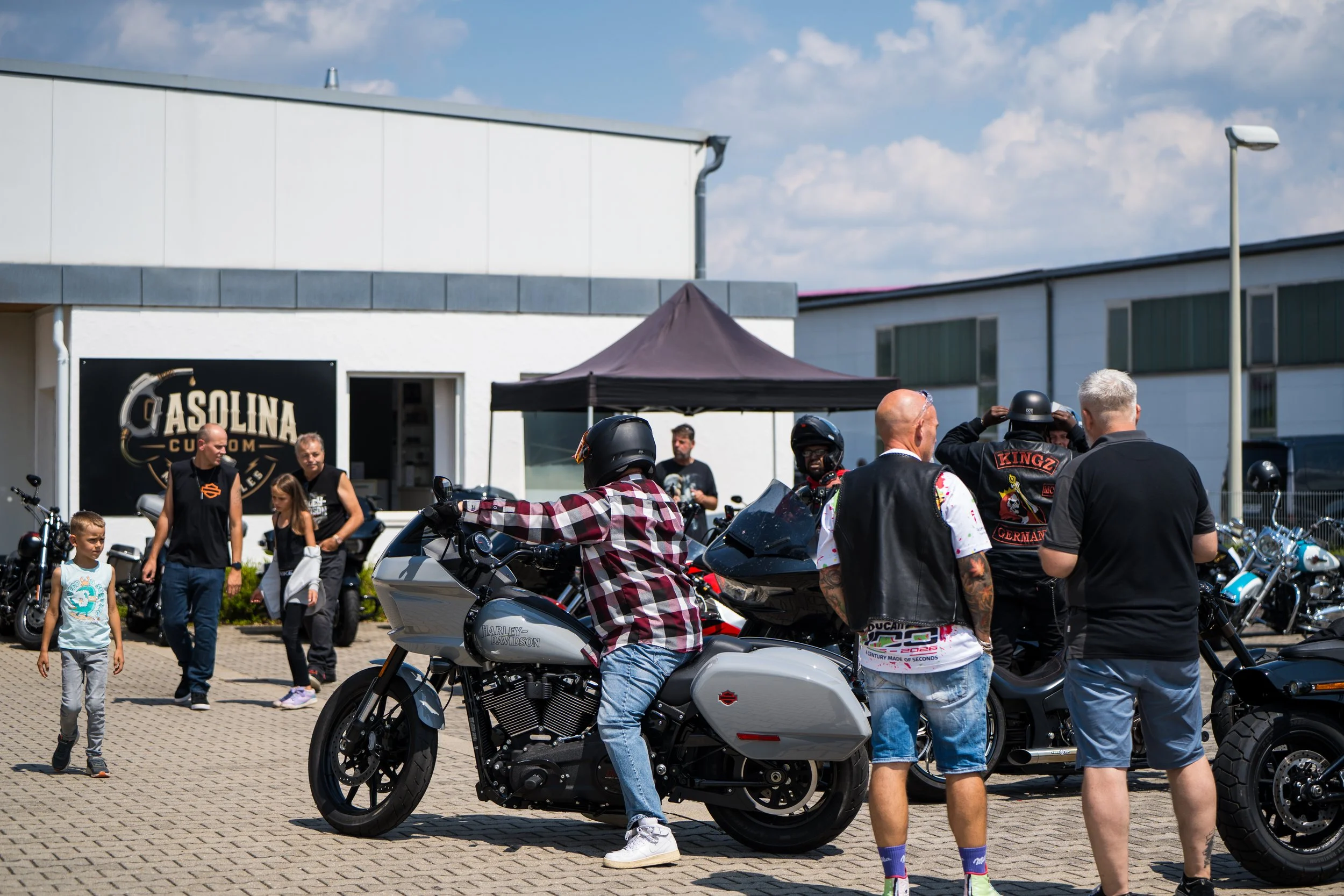 Menschen versammeln sich bei einer Motorradausstellung vor einem weißen Gebäude. Es sind verschiedene Motorradmodelle zu sehen, einige Leute tragen Helme, und es gibt einen Stand mit der Aufschrift 'Casolina Custom Motorcycles'.