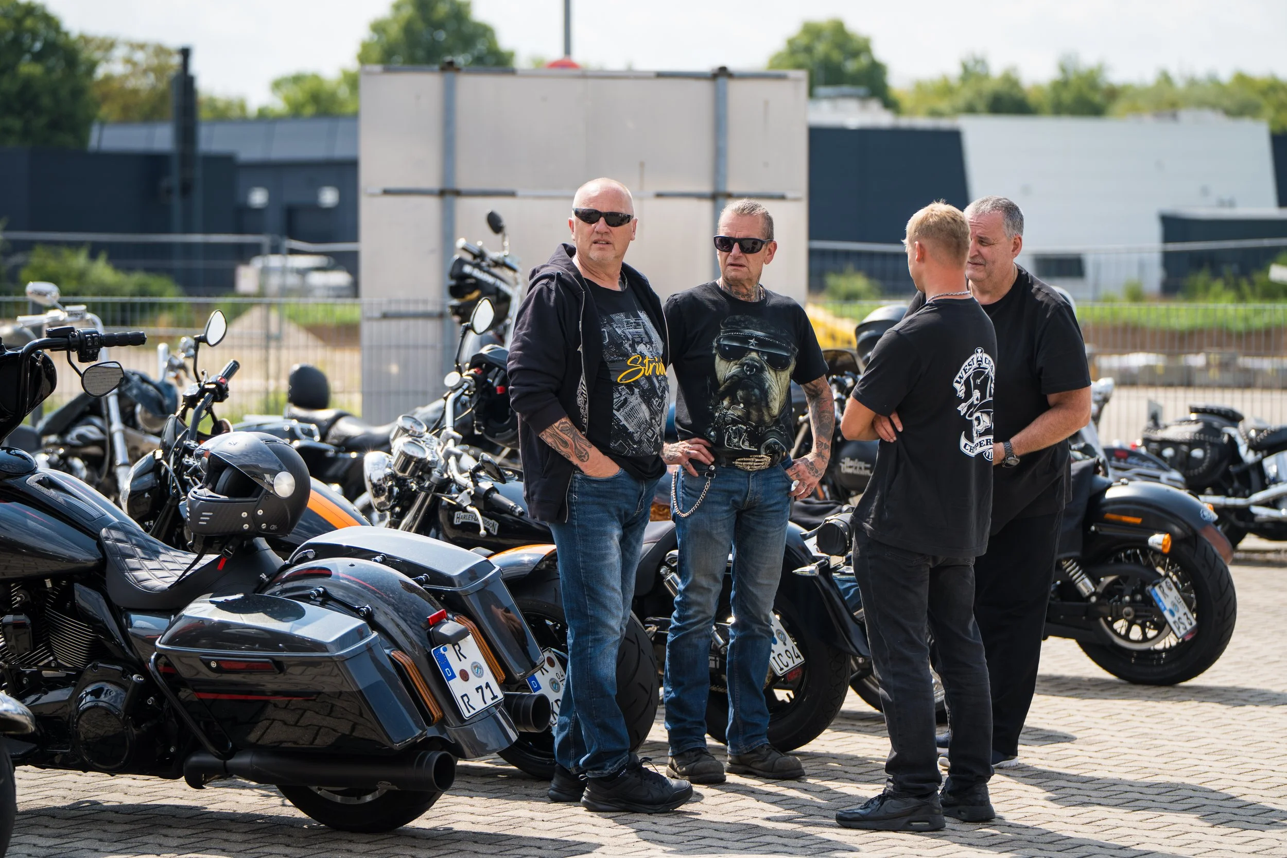 Eine Gruppe von vier Männern, die neben einer Reihe von Motorrädern in einem Parkplatz stehen und sich unterhalten, im Freien bei sonnigem Wetter. Bei der Eröffnungsfeier von Gasolina Custom Motorcycles.