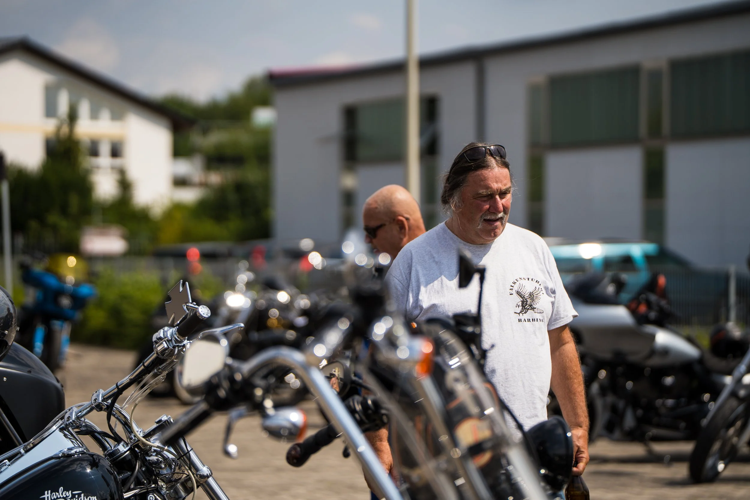 Ein Mann mit längeren Haaren und Sonnenbrille auf dem Kopf steht vor einer Reihe von Harley Davidson Motorrädern, im Hintergrund sind Häuser und bewölkter Himmel sichtbar.  Bei der Eröffnungsfeier von Gasolina Custom Motorcycles.
