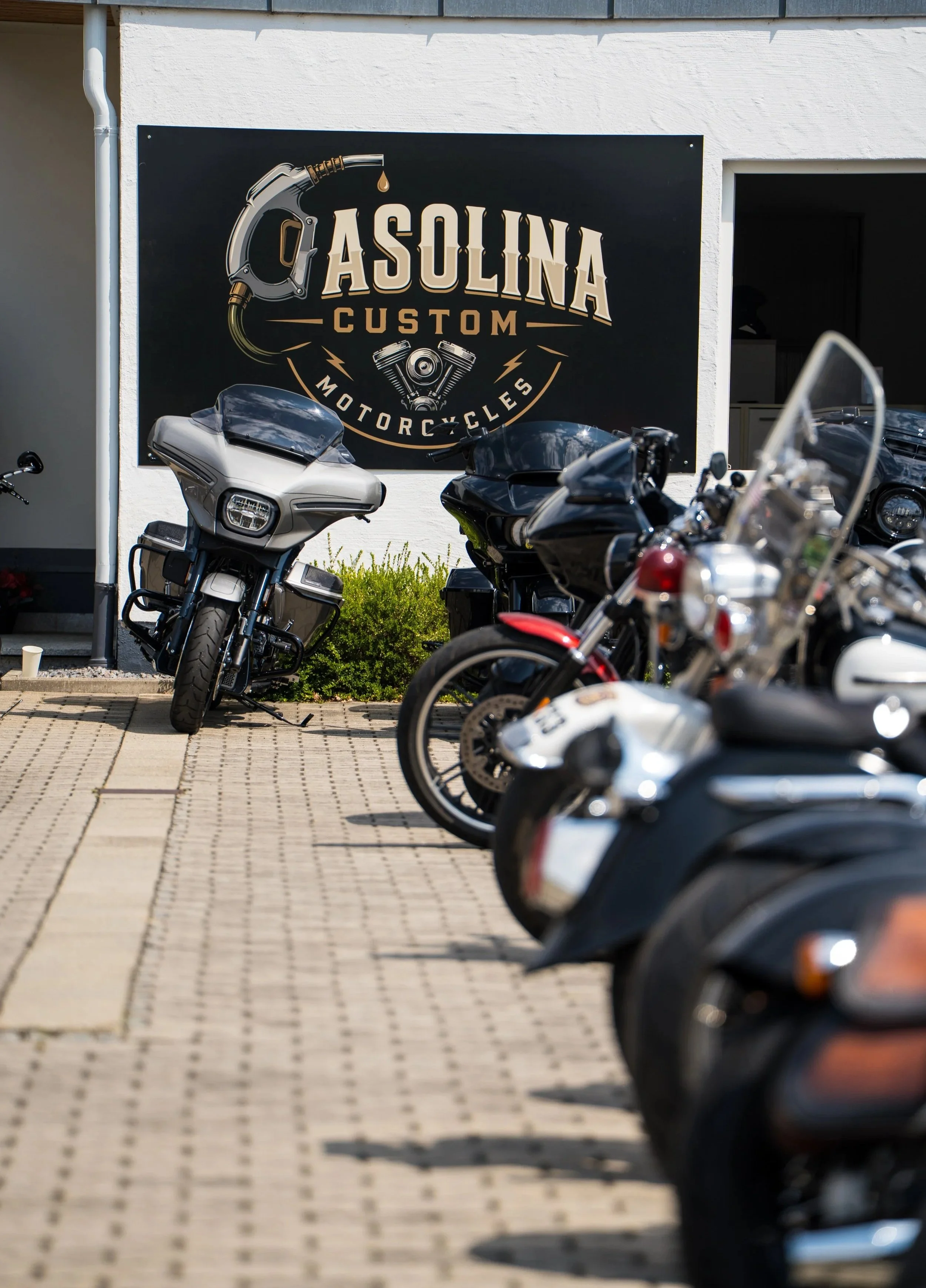 Mehrere Motorräder parken vor einem weißen Gebäude mit einem schwarzen Schilder, auf dem 'Asolina Custom Motorcycles' steht, und einem Werkzeug-Logo.