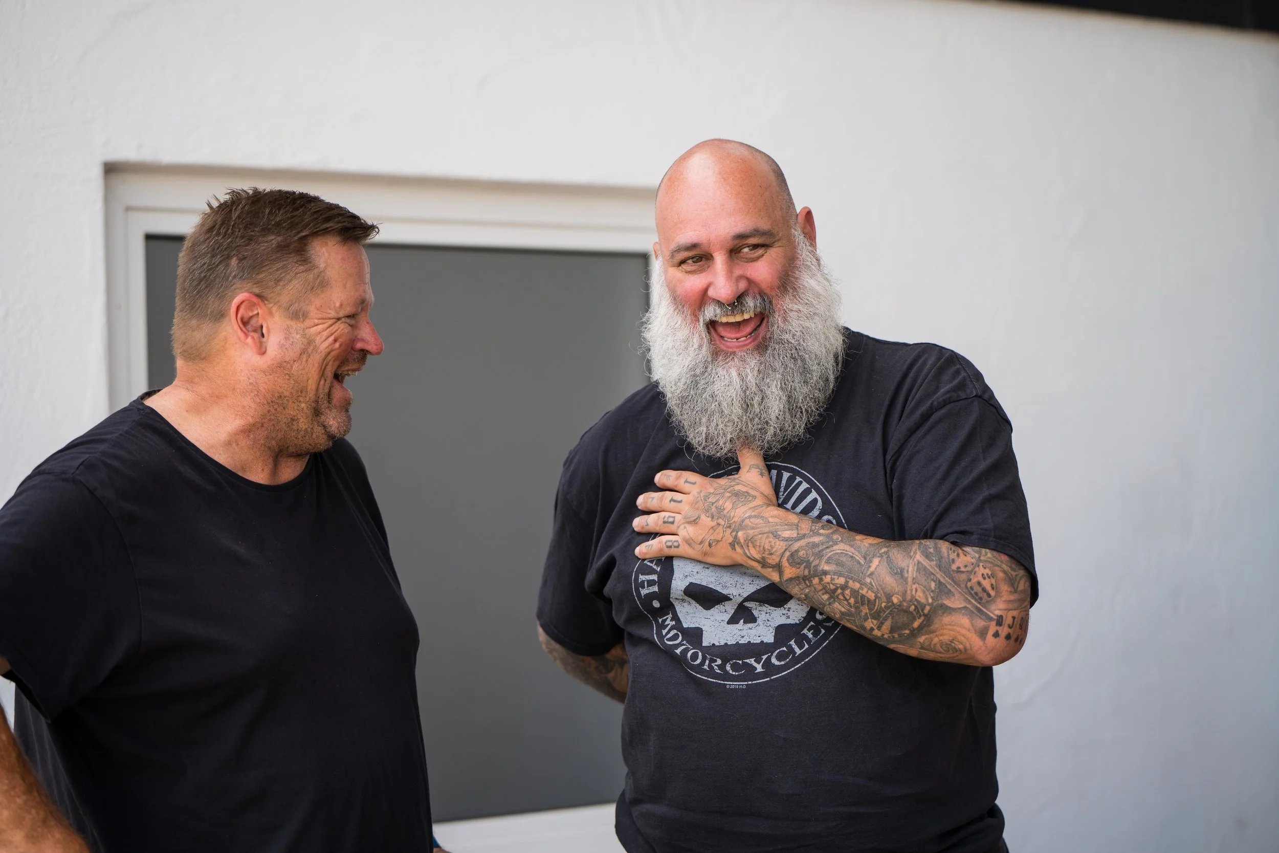 Zwei Männer lachen und reden miteinander, einer hat einen vollen grauen Bart und Tattoos am Arm, trägt ein schwarzes T-Shirt, der andere hat kurze Haare und träg ein schwarzes T-Shirt.   Bei der Eröffnungsfeier von Gasolina Custom Motorcycles.