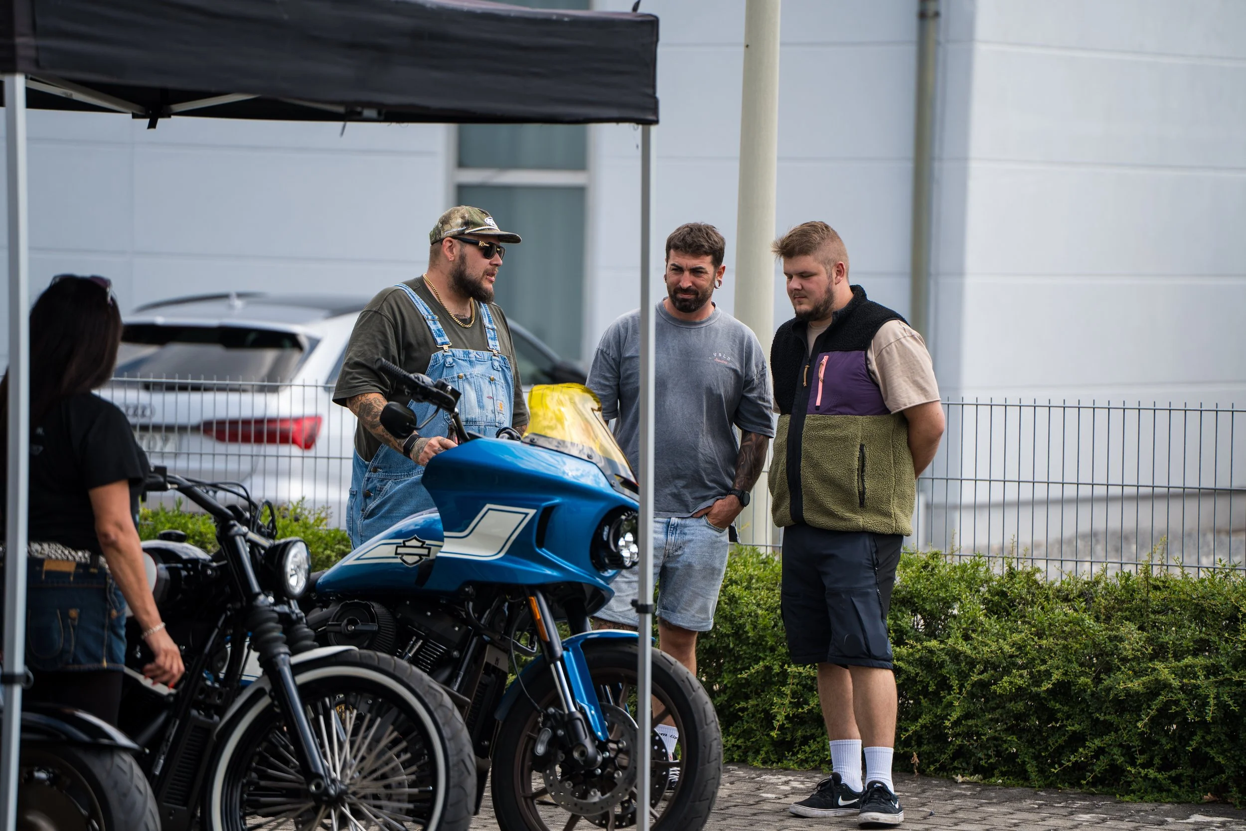 Drei Männer stehen hinter Motorrädern und unter einem Regenschirm im Freien.  Bei der Eröffnungsfeier von Gasolina Custom Motorcycles.