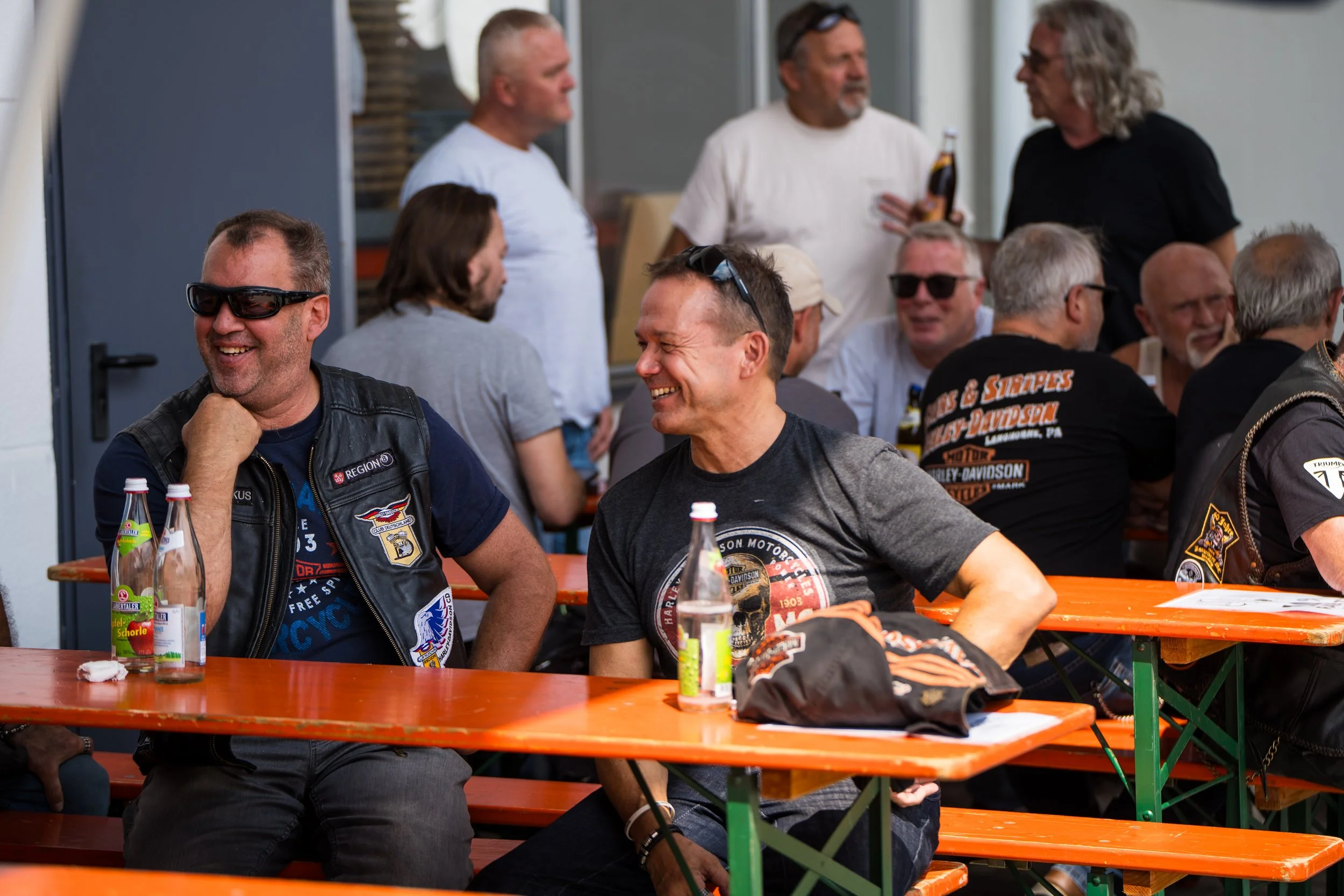 Menschen sitzen und stehen bei einer geselligen Zusammenkunft im Freien, lachen und unterhalten sich. Es gibt Getränke auf den Tischen, und die Stimmung ist fröhlich und entspannt.  Bei der Eröffnungsfeier von Gasolina Custom Motorcycles.