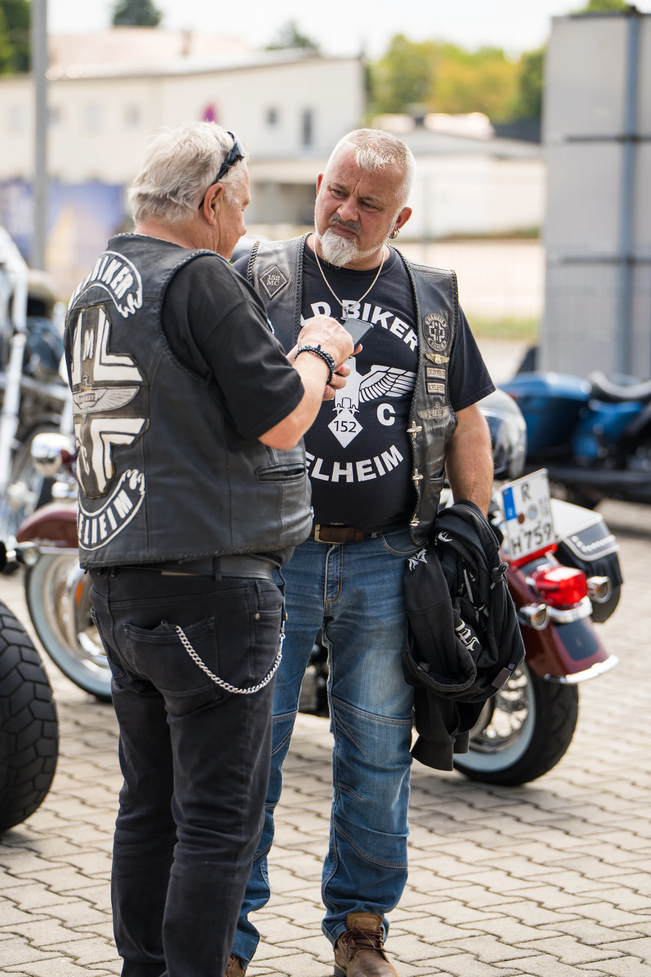 Zwei Männer, die sich unterhalten, stehen vor Motorrädern auf gepflastertem Boden. Sie tragen Lederjacken mit Biker-Logos, blickt auf Handy und trägt Schmuck.  Bei der Eröffnungsfeier von Gasolina Custom Motorcycles.