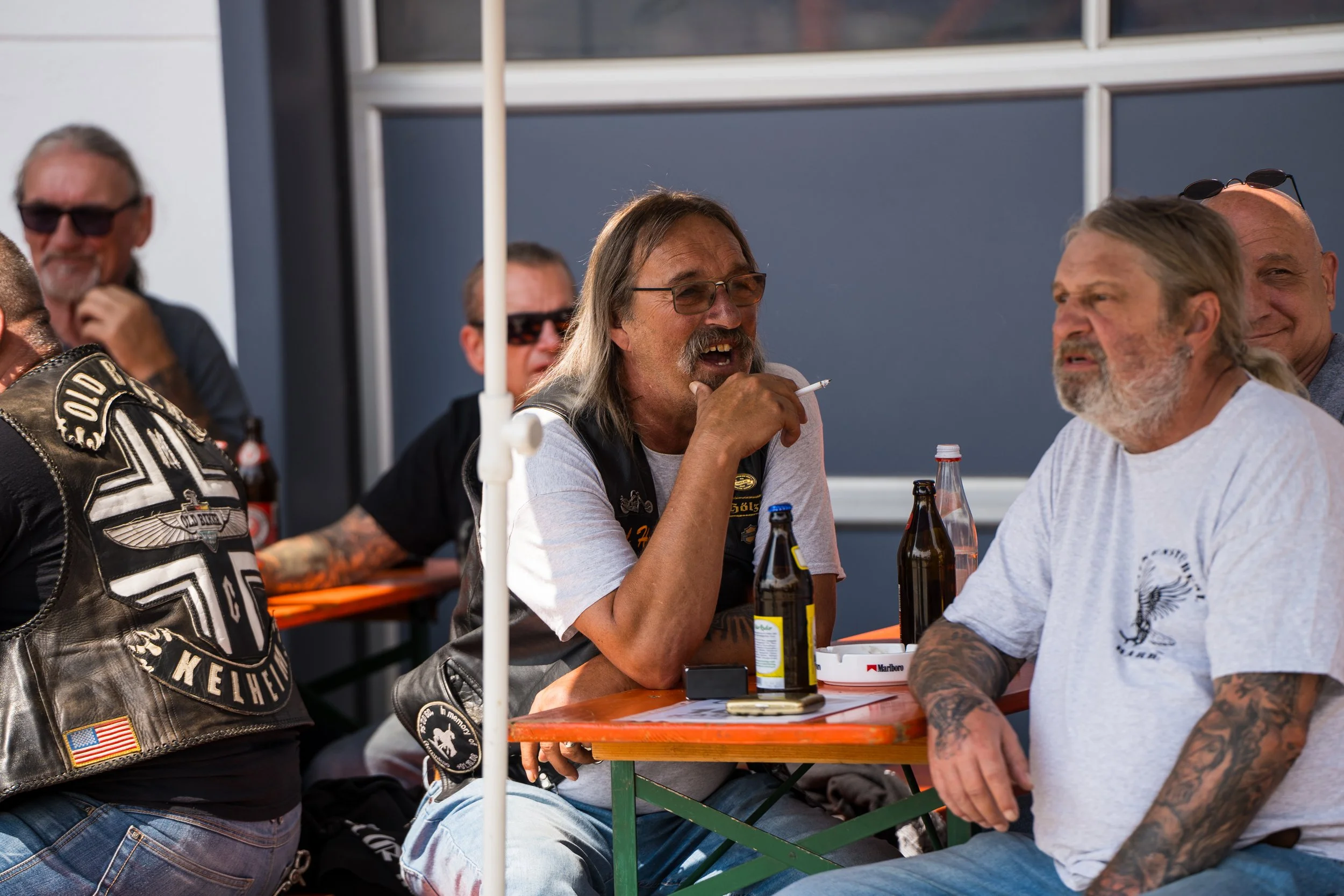 Gruppe von Motorradfahrern im Gespräch an einem Tisch im Freien, mit Bierflaschen auf dem Tisch.  Bei der Eröffnungsfeier von Gasolina Custom Motorcycles.