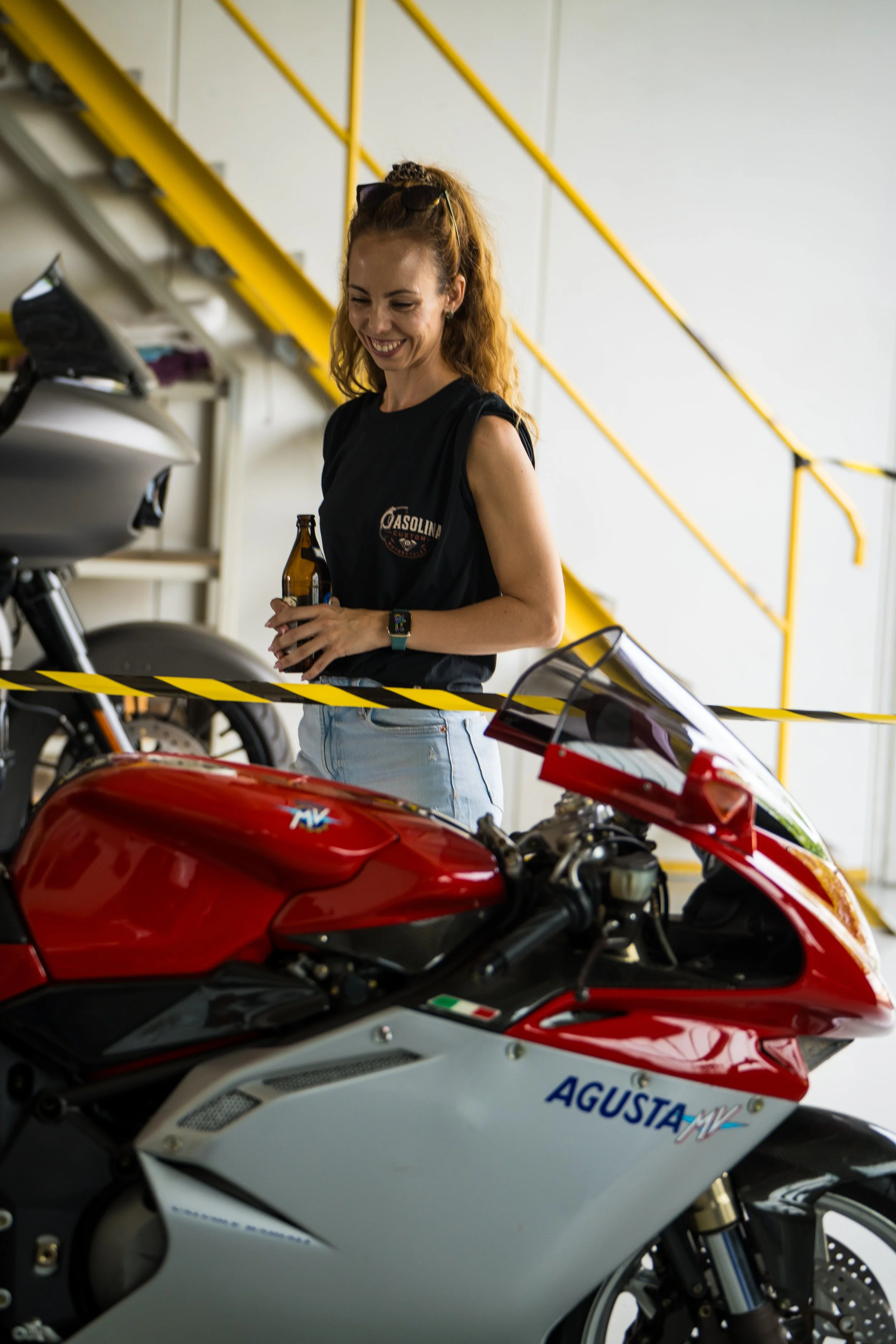 Eine lachende Frau mit roten Locken, Sonnenbrille auf dem Kopf, hält ein Bier und steht neben roten und weißen Motorrädern in einer Garage mit gelben Geländern. Bei der Eröffnungsfeier von Gasolina Custom Motorcycles.