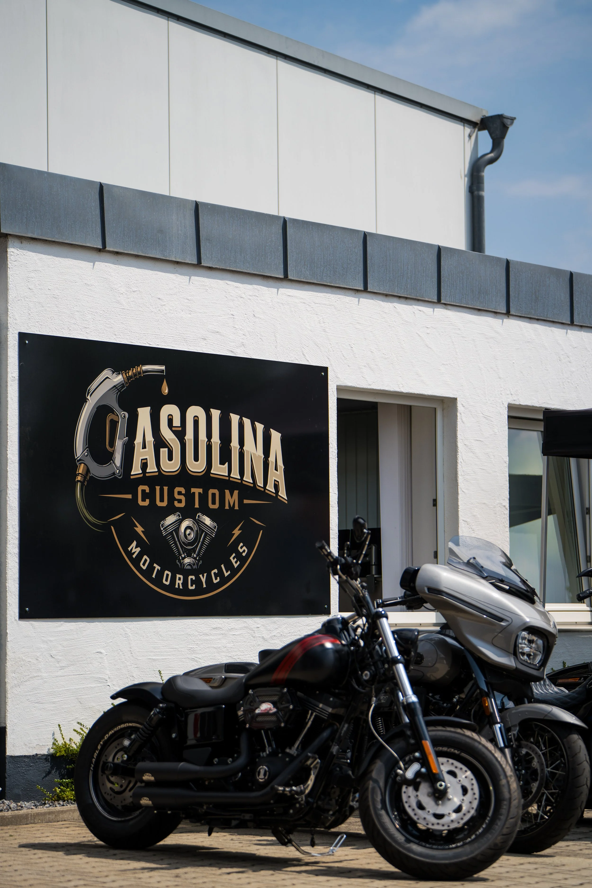 Motorräder vor einem Gebäude mit einem schwarzen Schild, das das Gasolina Custom Motorcycles-Logo zeigt, bei sonnigem Wetter. Bei der Eröffnungsfeier von Gasolina Custom Motorcycles.