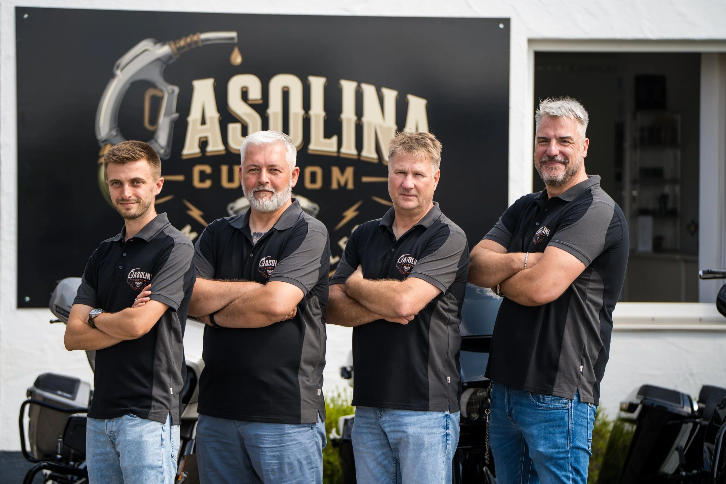 Vier Männer stehen vor einem schwarzen Firmenlogo an der Außenwand eines Gebäudes, tragen schwarze Polohemden mit Firmenlogo, Arme verschränkt. Bei der Eröffnungsfeier von Gasolina Custom Motorcycles.