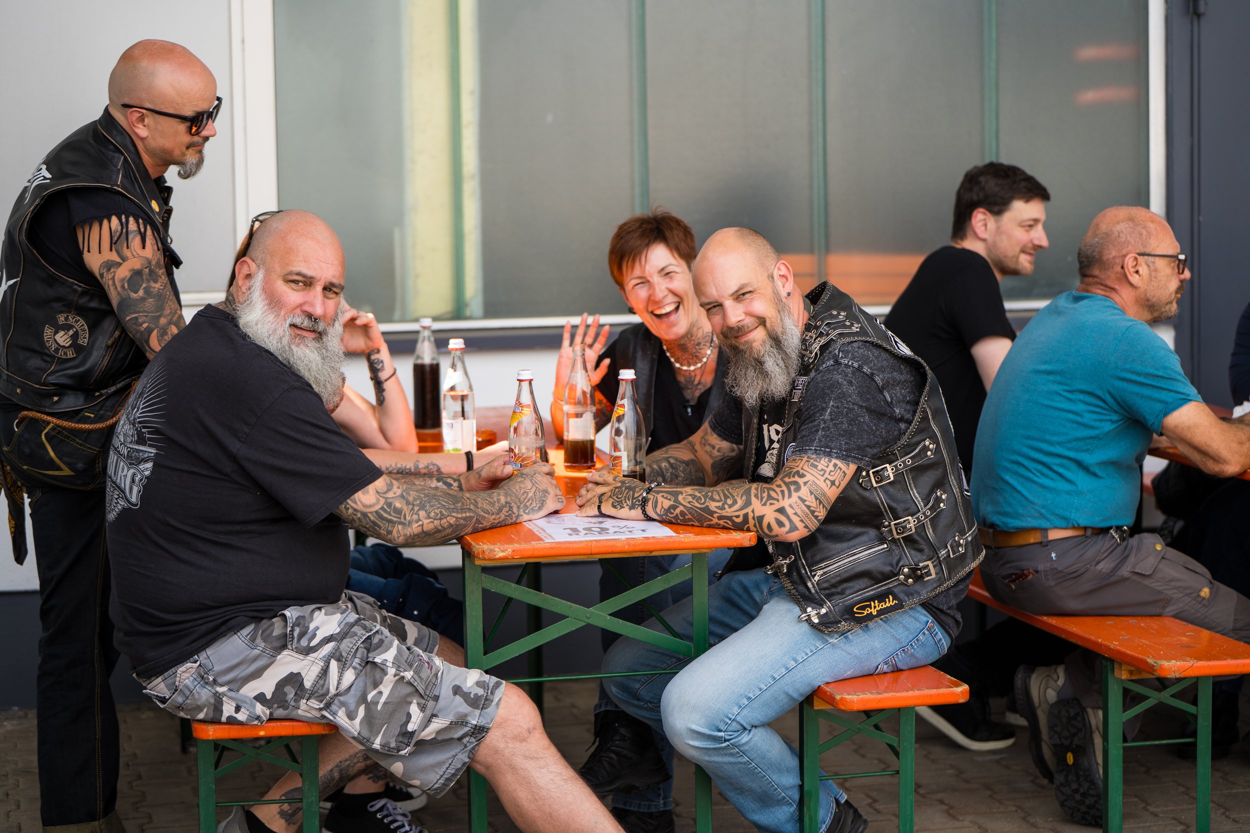 Gruppe von Menschen, überwiegend Männer, sitzt an einem langen Tisch im Freien. Sie lachen, trinken Cola und tragen meist dunkle Kleidung mit Lederjacken.  Bei der Eröffnungsfeier von Gasolina Custom Motorcycles.