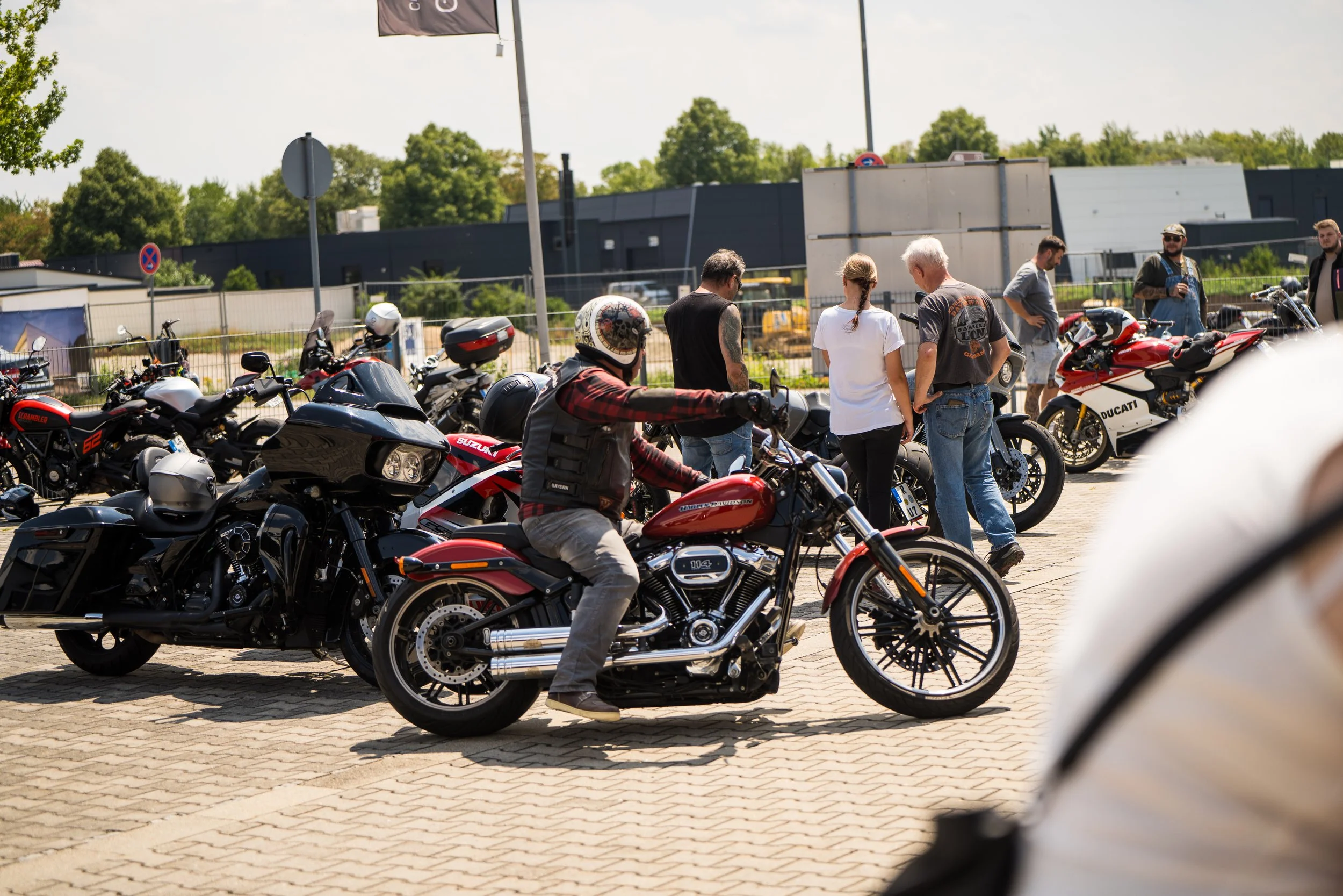 Mehrere Personen stehen neben und auf Motorrädern. Manche tragen Helme und Motorradkleidung, während andere casual gekleidet sind. Es sind verschiedene Motorräder, darunter eine Ducati.  Bei der Eröffnungsfeier von Gasolina Custom Motorcycles.