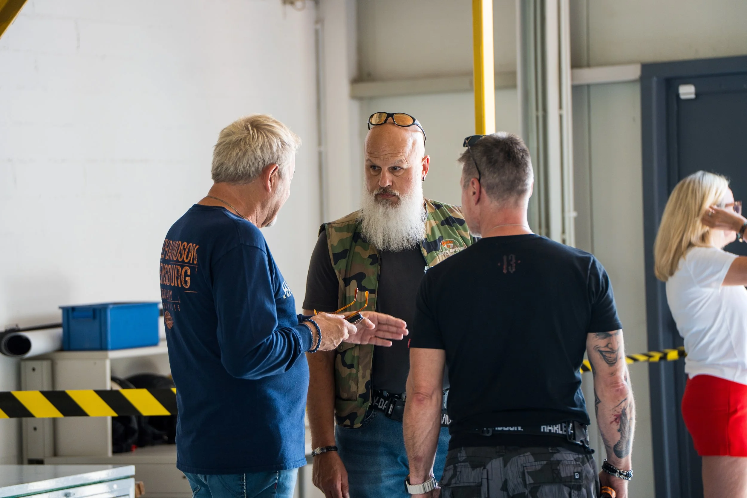 Drei Männer in einem Gespräch in einem Industriegebäude, einer davon trägt eine bewaffnete Weste, eine Frau steht im Hintergrund.  Bei der Eröffnungsfeier von Gasolina Custom Motorcycles.