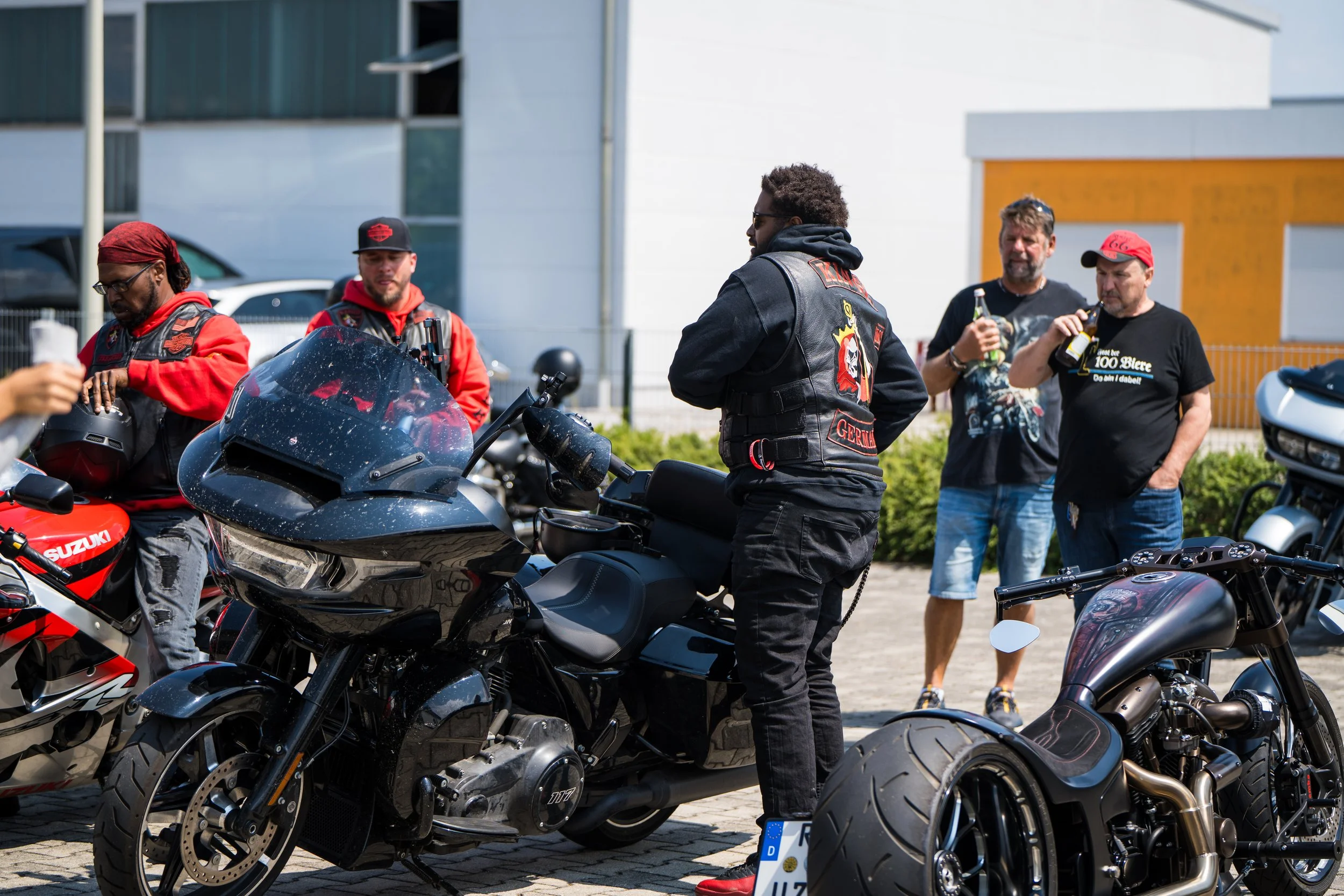Ein Männergruppe bei einem Treffen mit Motorrädern in einer urbanen Umgebung, einige Männer trinken Bier, während andere mit Motorrädern und Schutzkleidung unterwegs sind. Bei der Eröffnungsfeier von Gasolina Custom Motorcycles.