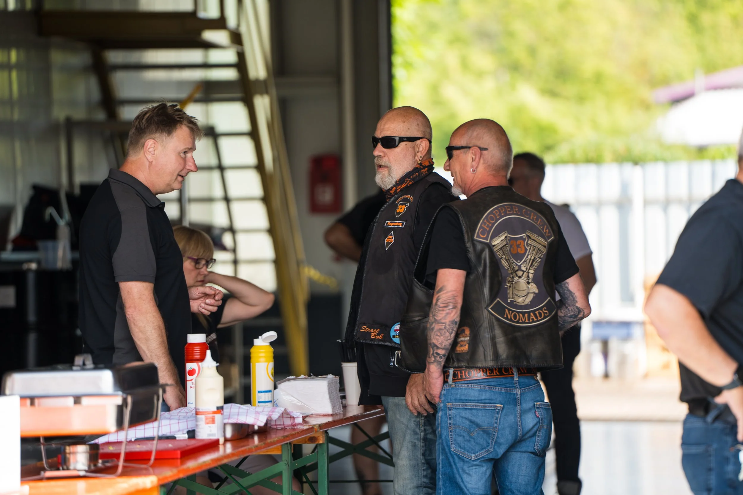 Biker unterhält sich mit einem Mann an einem Stand bei einer Veranstaltung im Innenbereich mit mehreren Personen, Bierzeltgarnituren und Grün im Hintergrund.  Bei der Eröffnungsfeier von Gasolina Custom Motorcycles.