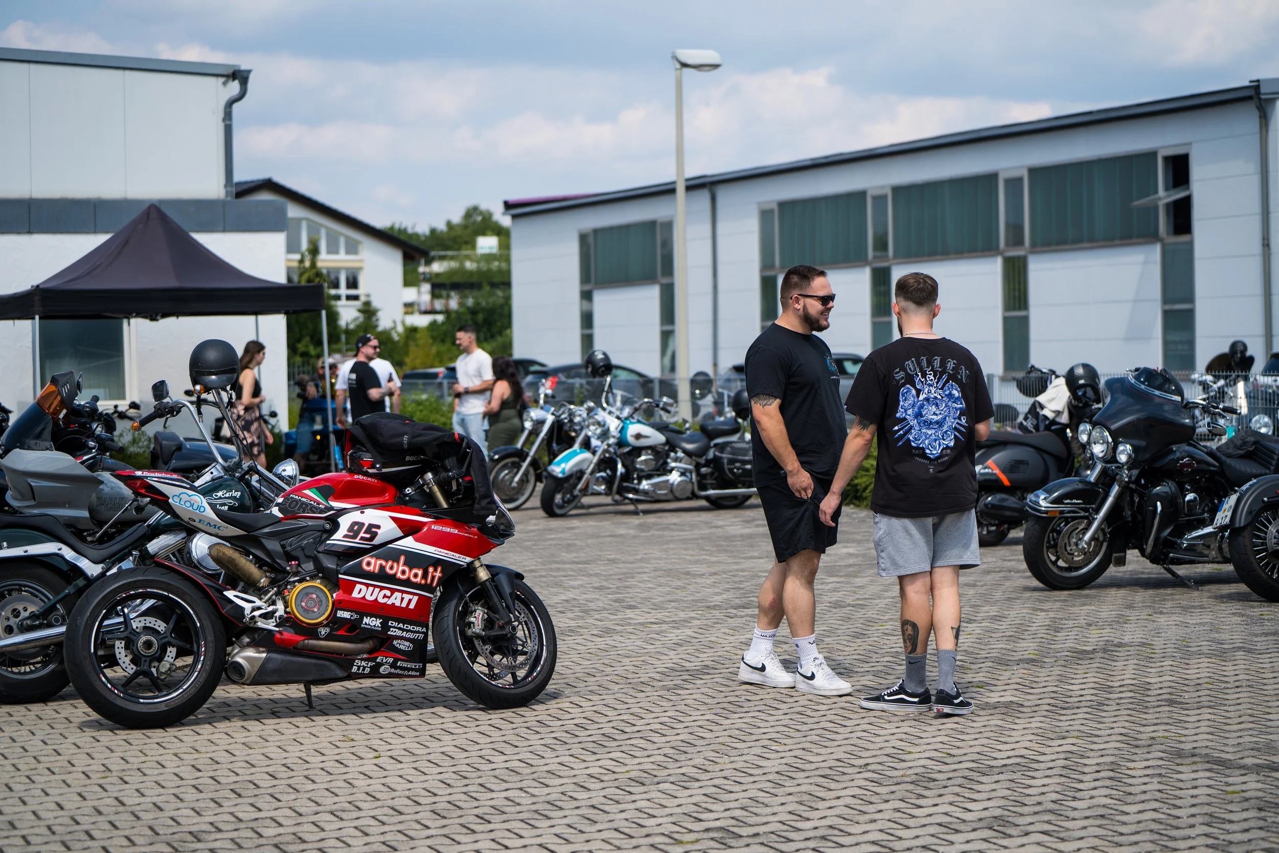 Menschen stehen neben Motorrädern und sprechen im Freien vor modernen Gebäuden. Bei der Eröffnungsfeier von Gasolina Custom Motorcycles.