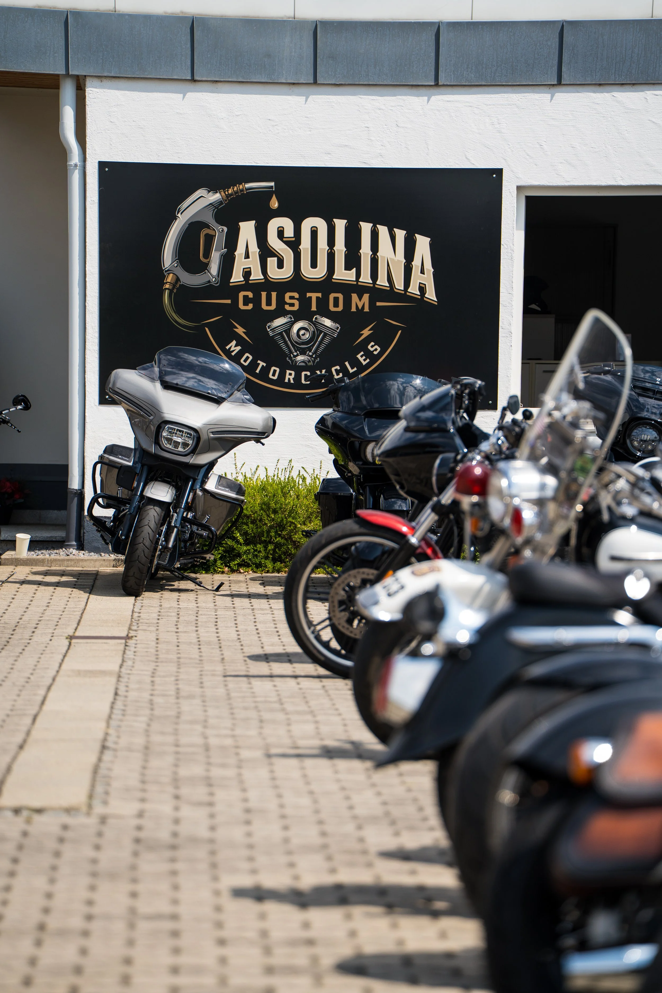 Mehrere Motorräder parken vor einem Gebäude mit einem schwarzen Schild, das 'GASOLINA CUSTOM MOTORCYCLES' zeigt.