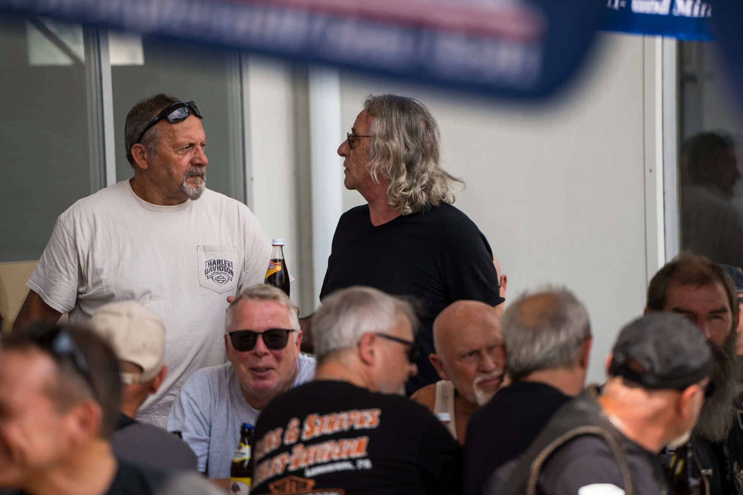 Zwei Männer im Gespräch, während ein großer Menschenansammlung unter ihnen ist.  Bei der Eröffnungsfeier von Gasolina Custom Motorcycles.