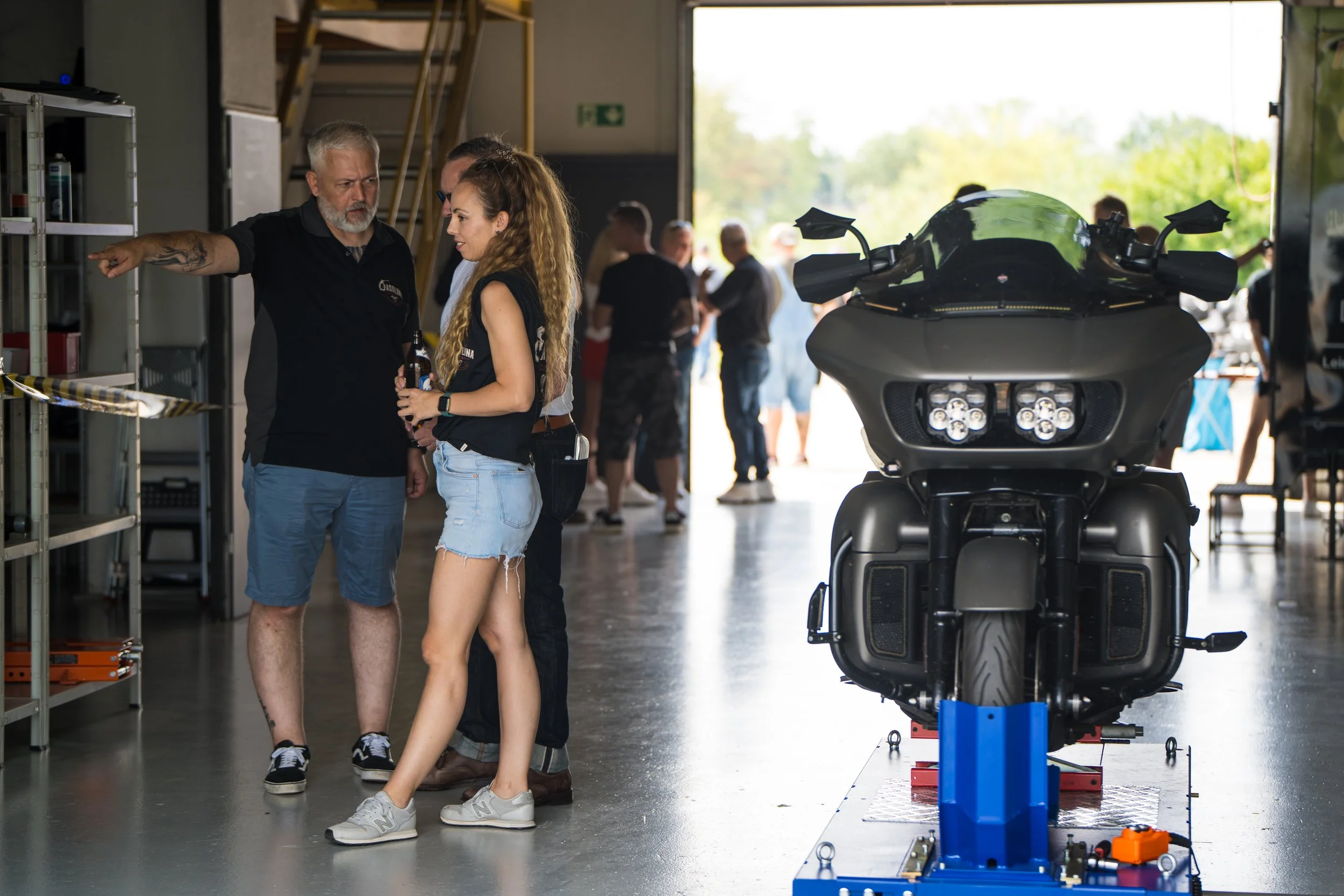 Menschen in einer Garage mit einem Motorrad und technischen Werkzeugen, im Hintergrund andere Personen und ein offener Torbereich bei Tageslicht. Bei der Eröffnungsfeier von Gasolina Custom Motorcycles.