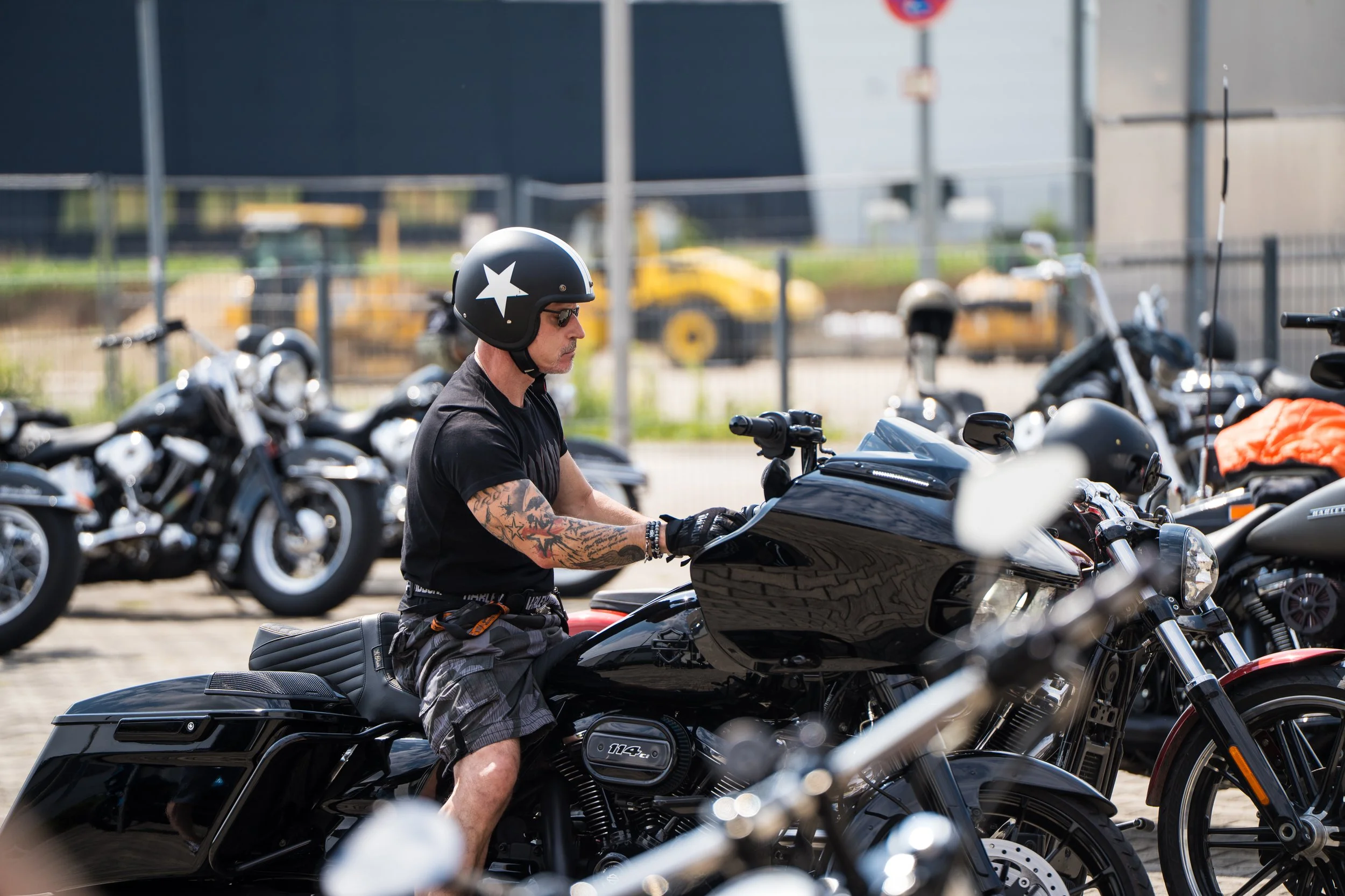 Ein Mann mit Tattoos und Sonnenbrille sitzt auf einem schwarzen Motorrad, umgeben von weiteren Motorrädern,  Bei der Eröffnungsfeier von Gasolina Custom Motorcycles.