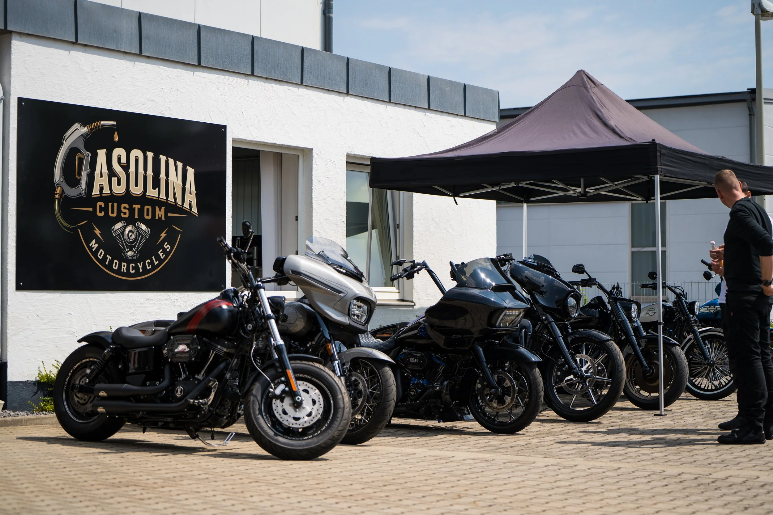 Mehrere Motorräder parken vor einem weißen Gebäude mit einem schwarzen Ausschank. Ein Mann steht neben den Motorrädern.  Bei der Eröffnungsfeier von Gasolina Custom Motorcycles.