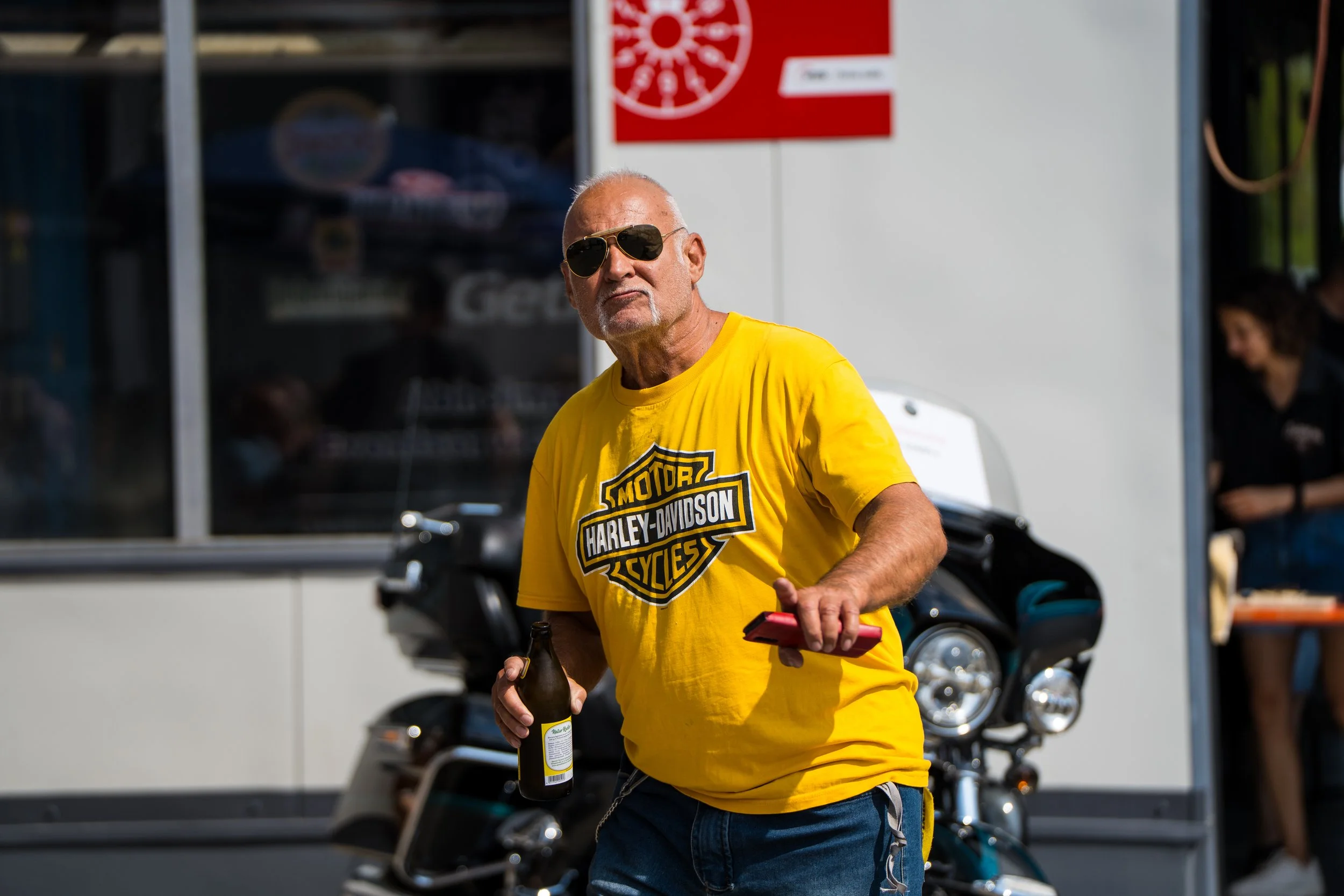 Ein älterer Mann trägt eine gelbe Harley-Davidson-T-Shirt, Sonnenbrille und hält ein Bier in der Hand, während er neben einem Motorrad steht. Bei der Eröffnungsfeier von Gasolina Custom Motorcycles.