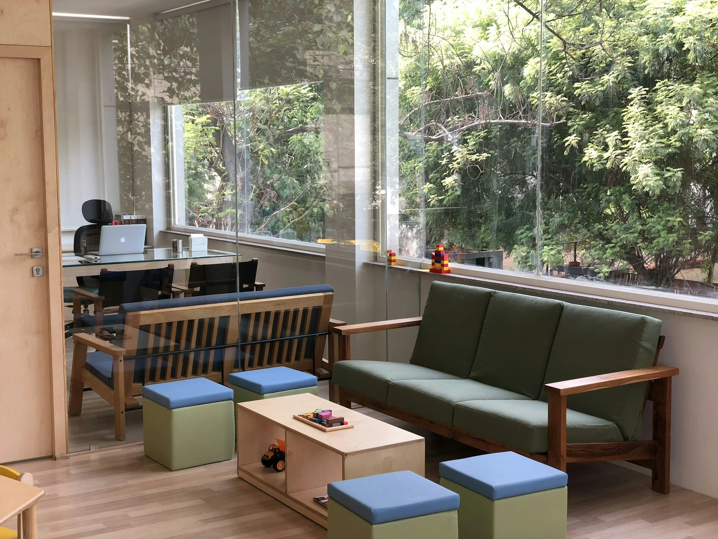 A modern office or classroom space with large windows showing green trees outside. The room contains a desk with a MacBook laptop, a chair, and shelves with various items. In the foreground, there is a green sofa, small blue stools, and a white table with toys on it.