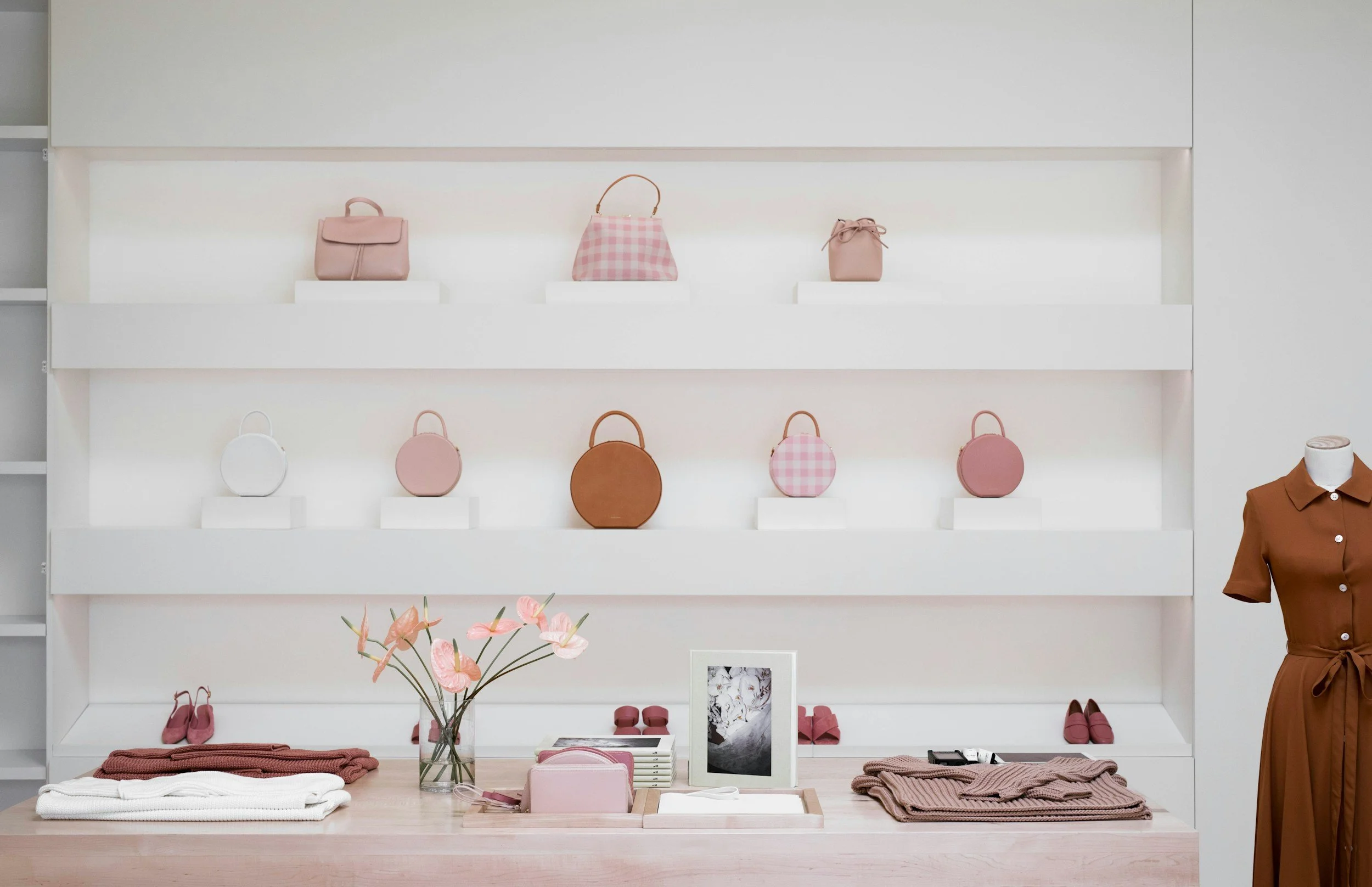Display of blush pink handbags on white shelves and a table with pink shoes, a pink purse, and pink flowers in a retail store.