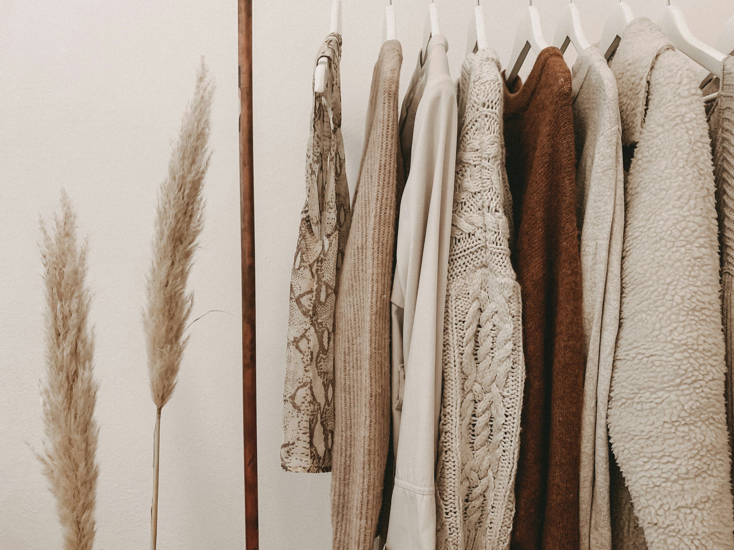 A clothing rack with various beige and brown sweaters and cardigans, and some woven and knit textures, next to pampas grass plumes.