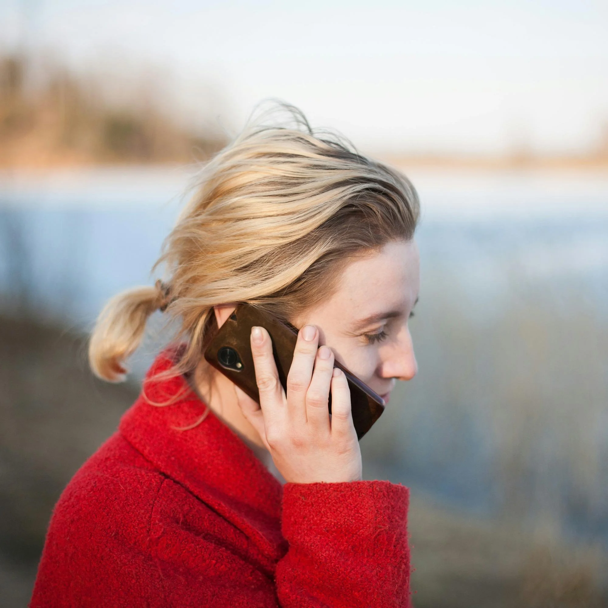 Close-up of a woman with blonde hair wearing a red coat and talking on the phone outside.