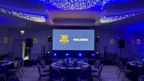 A conference room decorated for a 30-year anniversary celebration with a large screen displaying a welcome message, stage lights, round tables with place settings, and ceiling lights with patterned blue lighting.