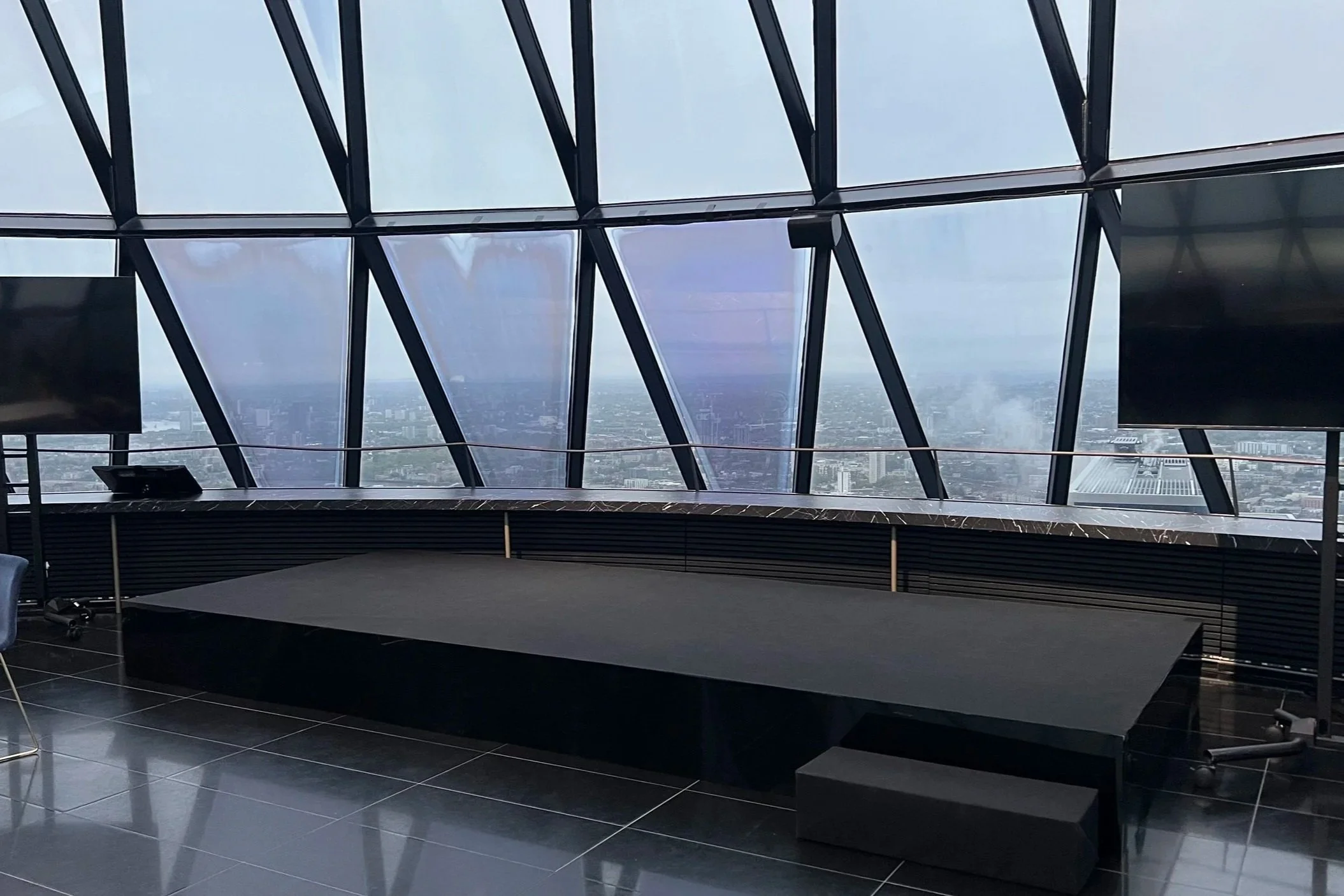 Interior of a modern building with large glass windows showing a cityscape view, a black stage with a raised platform, and TV screens mounted on the walls.