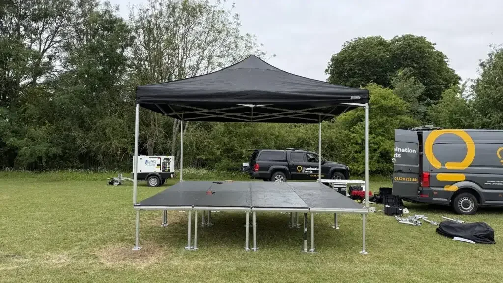 Stage setup in an open grassy area with a black canopy, elevated platform, and event equipment, including trucks and tools nearby.