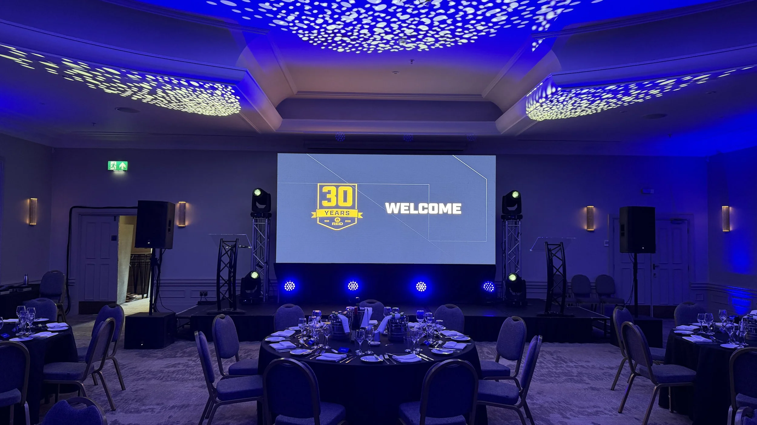 A conference room decorated for a 30-year anniversary celebration with a large screen displaying a welcome message, stage lights, round tables with place settings, and ceiling lights with patterned blue lighting.