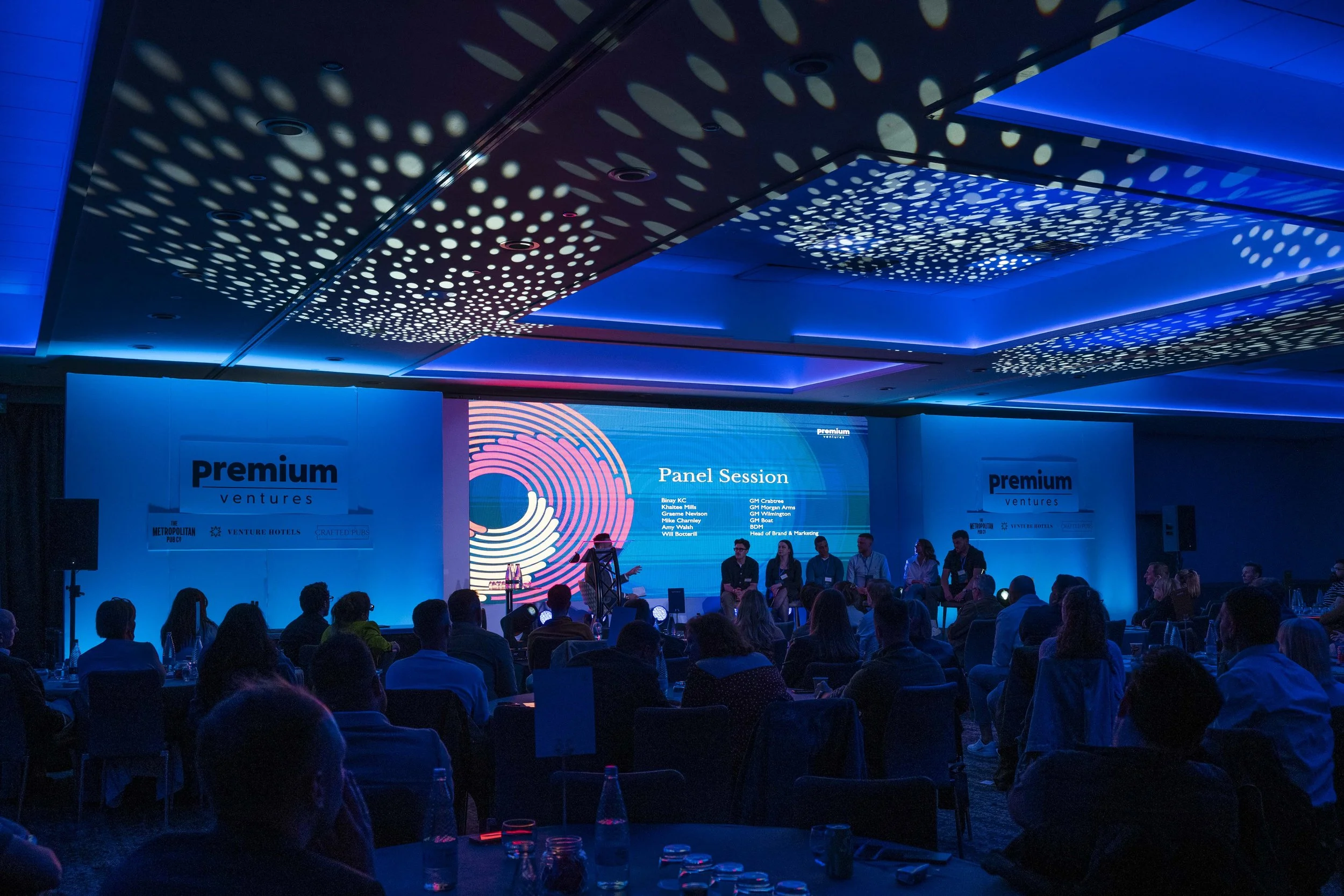 A conference room with a large screen displaying the words 'Panel Session' and a list of panelists' names. The room is filled with seated attendees, and the ceiling has decorative, illuminated circular patterns.