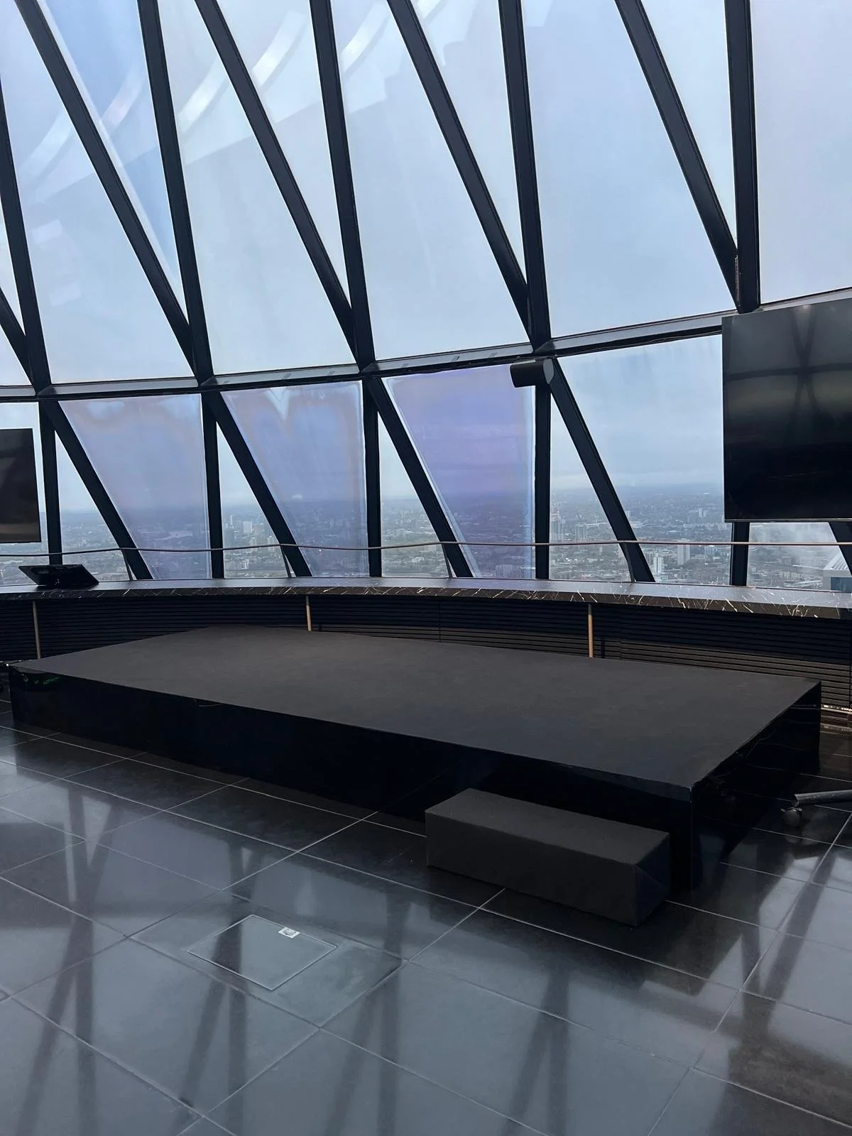Interior of a modern high-rise building with a black stage, a small black step, and large glass windows showing a city skyline.