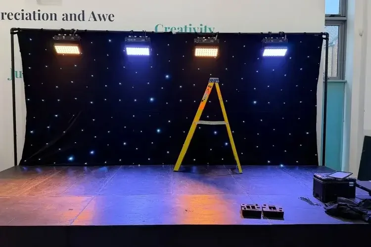 Stage with a black backdrop featuring small white lights, four stage lights mounted on top, a yellow ladder in the center, and equipment on the floor to the right.