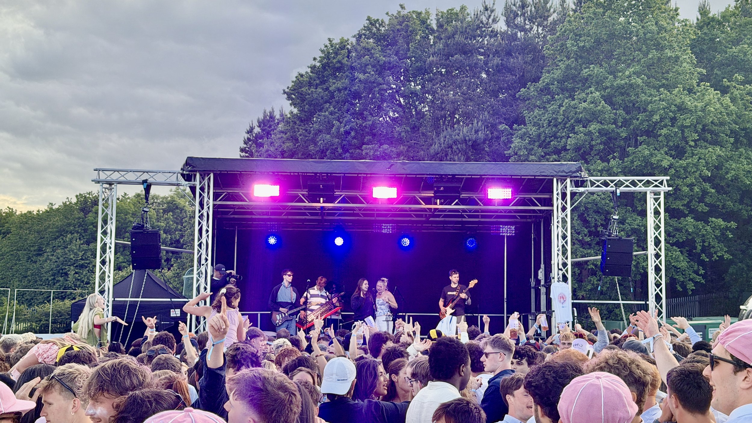 Outdoor concert with a band performing on stage under purple lights, crowd of people watching and raising hands, trees and cloudy sky in the background.