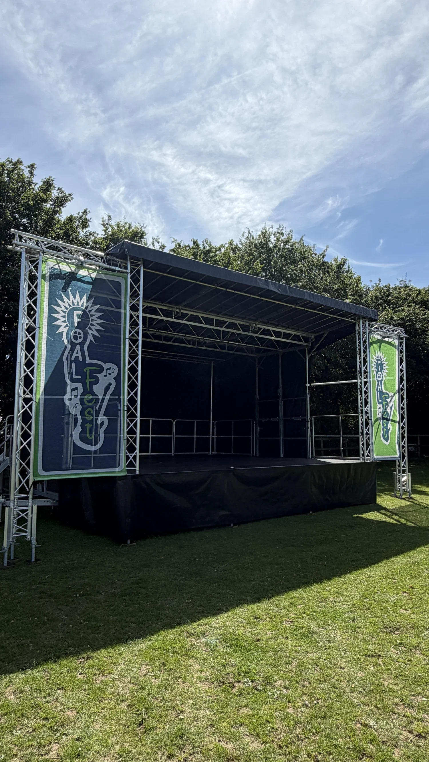 An empty outdoor stage with green and blue banners on each side reading 'Fall Fest,' set on a grassy field with trees and a partly cloudy sky in the background.