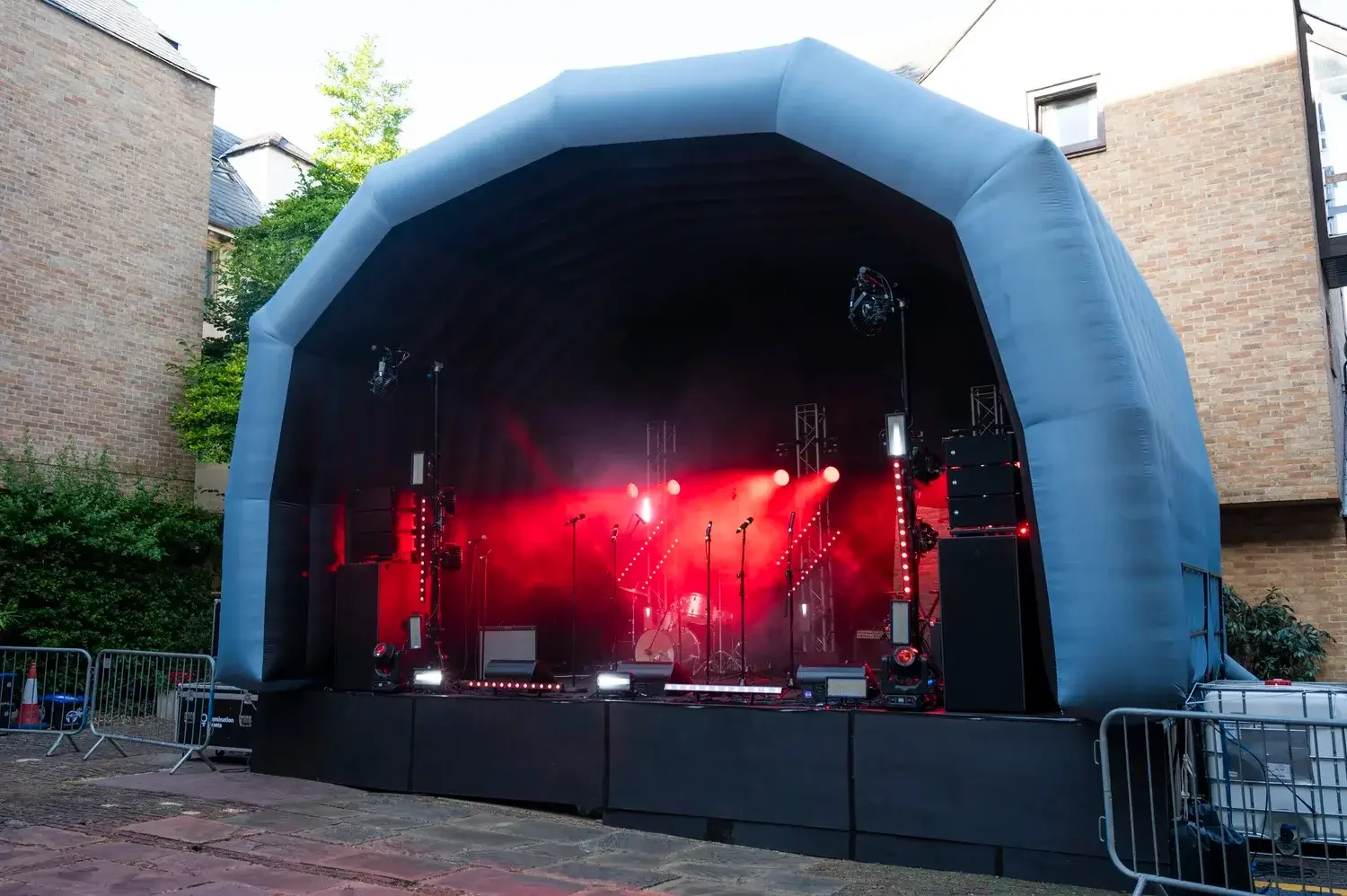 Empty outdoor stage with red lighting and fog effects, set up for a concert, surrounded by brick buildings and trees.