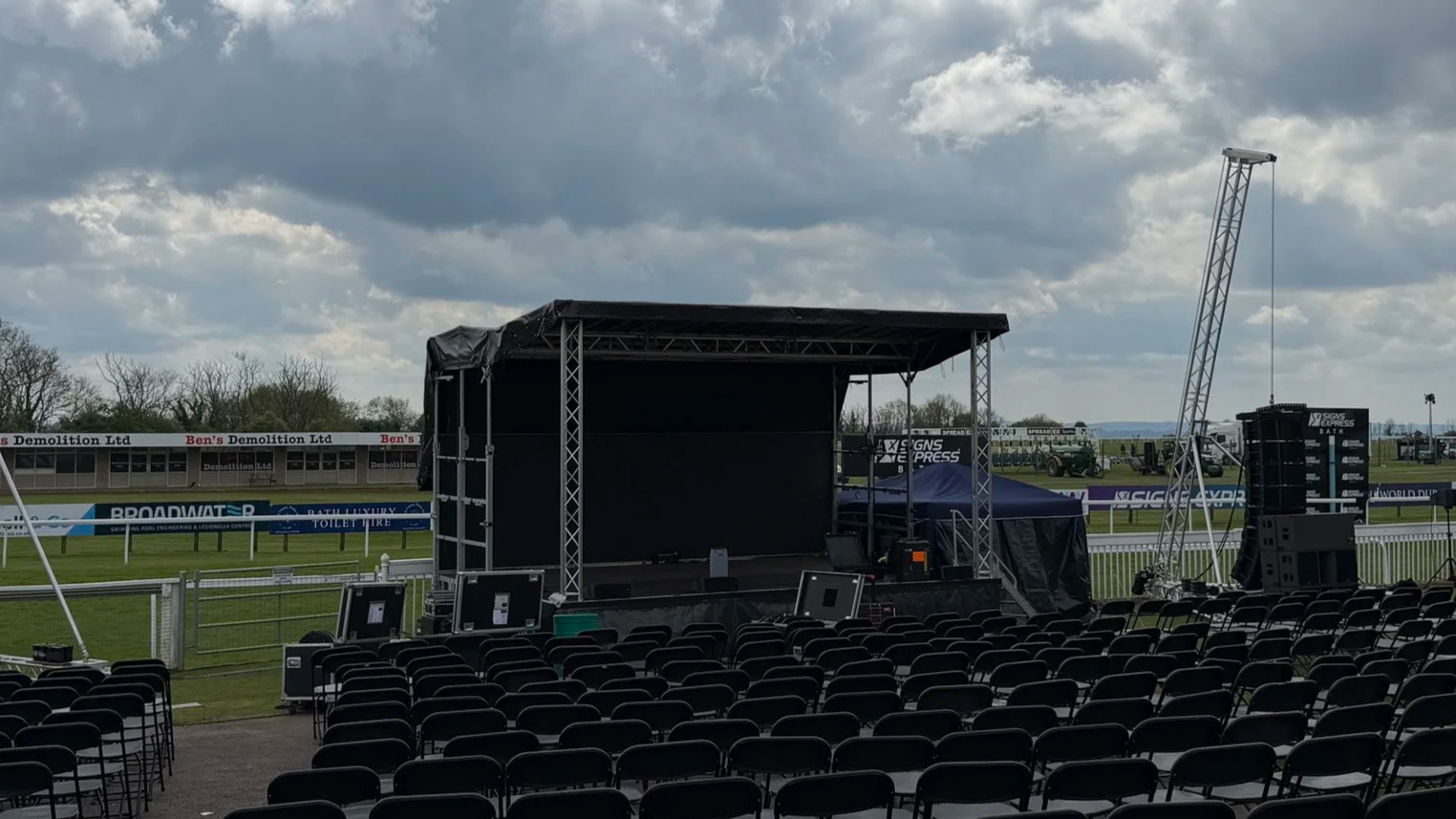 Empty outdoor stage setup with chairs facing a black stage, large speakers, and lighting equipment, under cloudy sky in a stadium or event field.