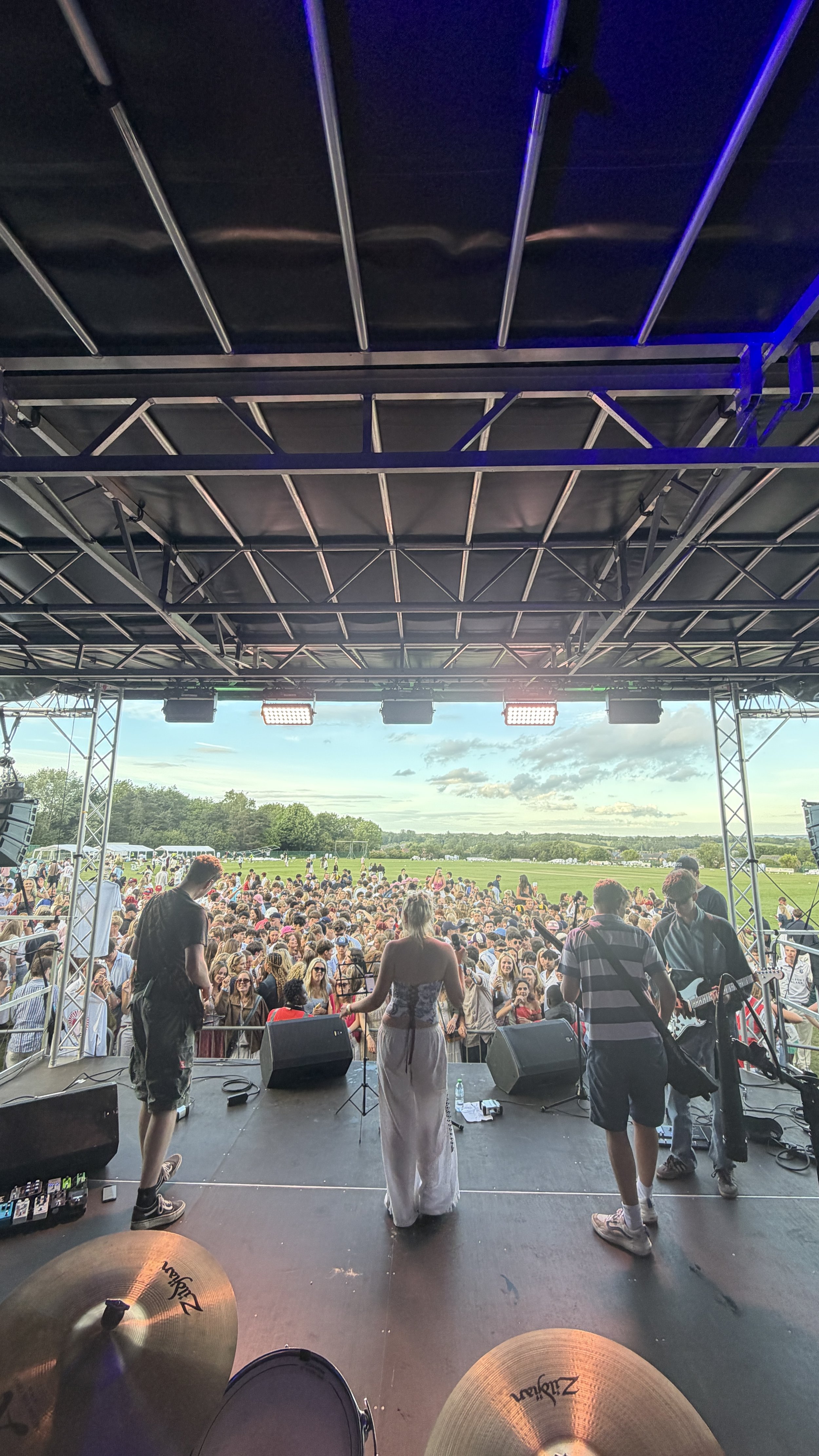 View from a stage overlooking a music concert with many people outdoors on a field, sky clear with some clouds, and a band on stage preparing to perform.