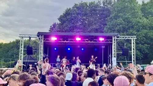 Outdoor concert with a band performing on stage under purple lights, crowd of people watching and raising hands, trees and cloudy sky in the background.
