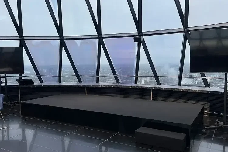 Interior of a modern building with large glass windows showing a cityscape view, a black stage with a raised platform, and TV screens mounted on the walls.