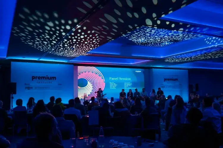 A conference room with a large screen displaying the words 'Panel Session' and a list of panelists' names. The room is filled with seated attendees, and the ceiling has decorative, illuminated circular patterns.