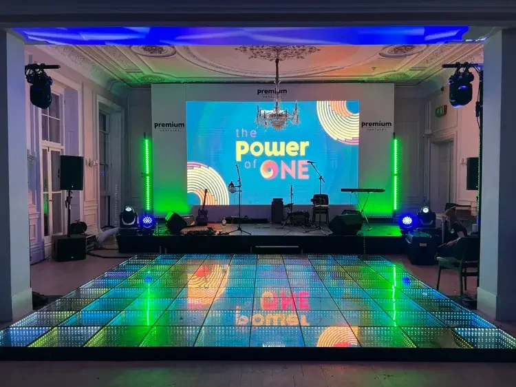 Event stage with a large LED screen displaying the text 'the power of ONE,' surrounded by colorful lights and musical instruments in a decorated room with a chandelier.