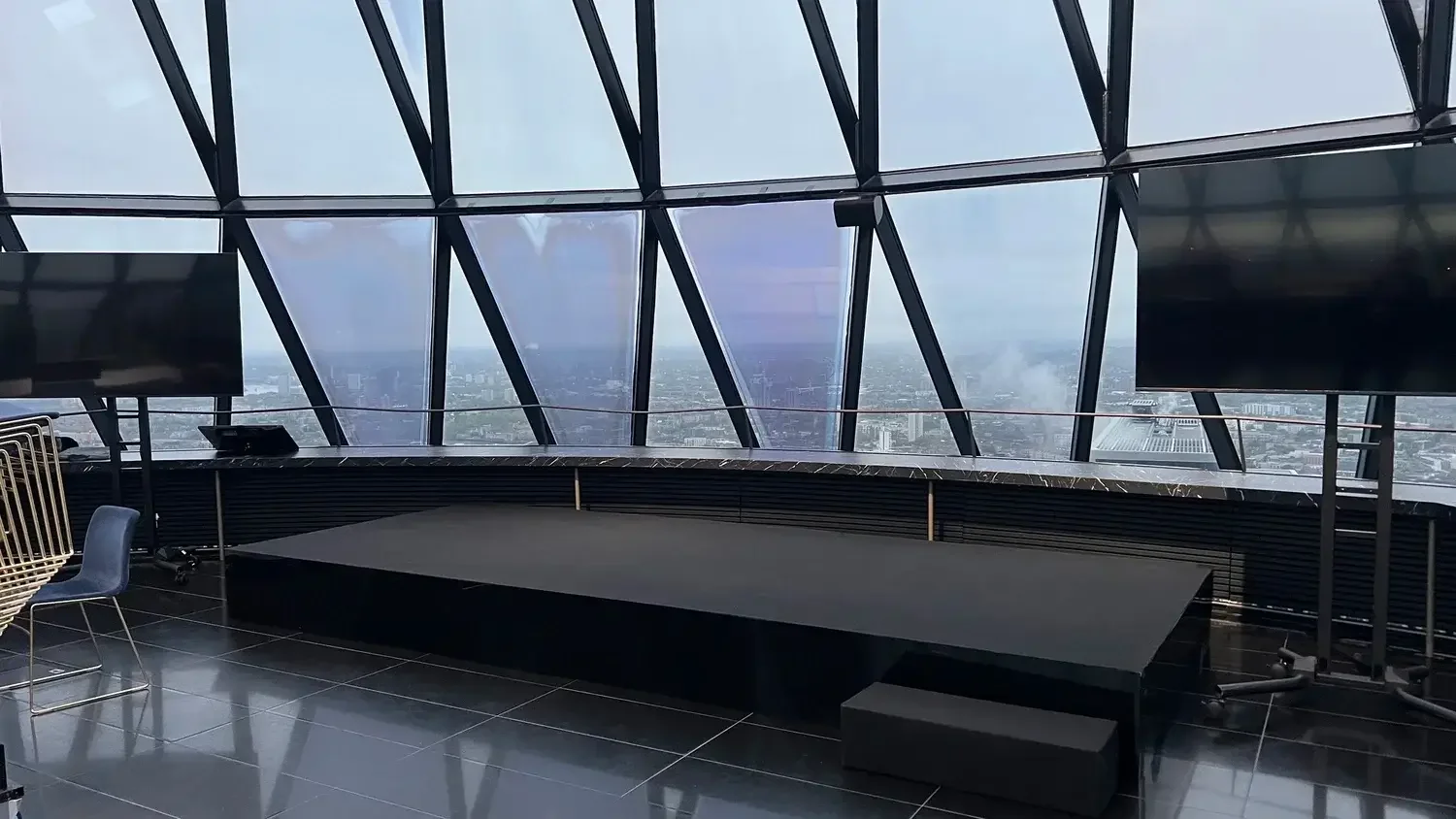 Interior view of a modern building with large glass windows showing a cityscape, a black marble counter, and TV screens, with chairs nearby.
