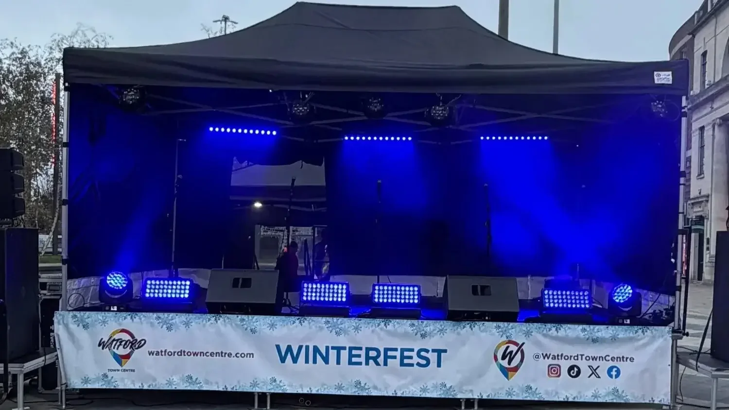 A stage setup for the Watford Winterfest event featuring blue stage lighting, speakers, microphones, and a banner with event details and social media icons in an outdoor urban setting.