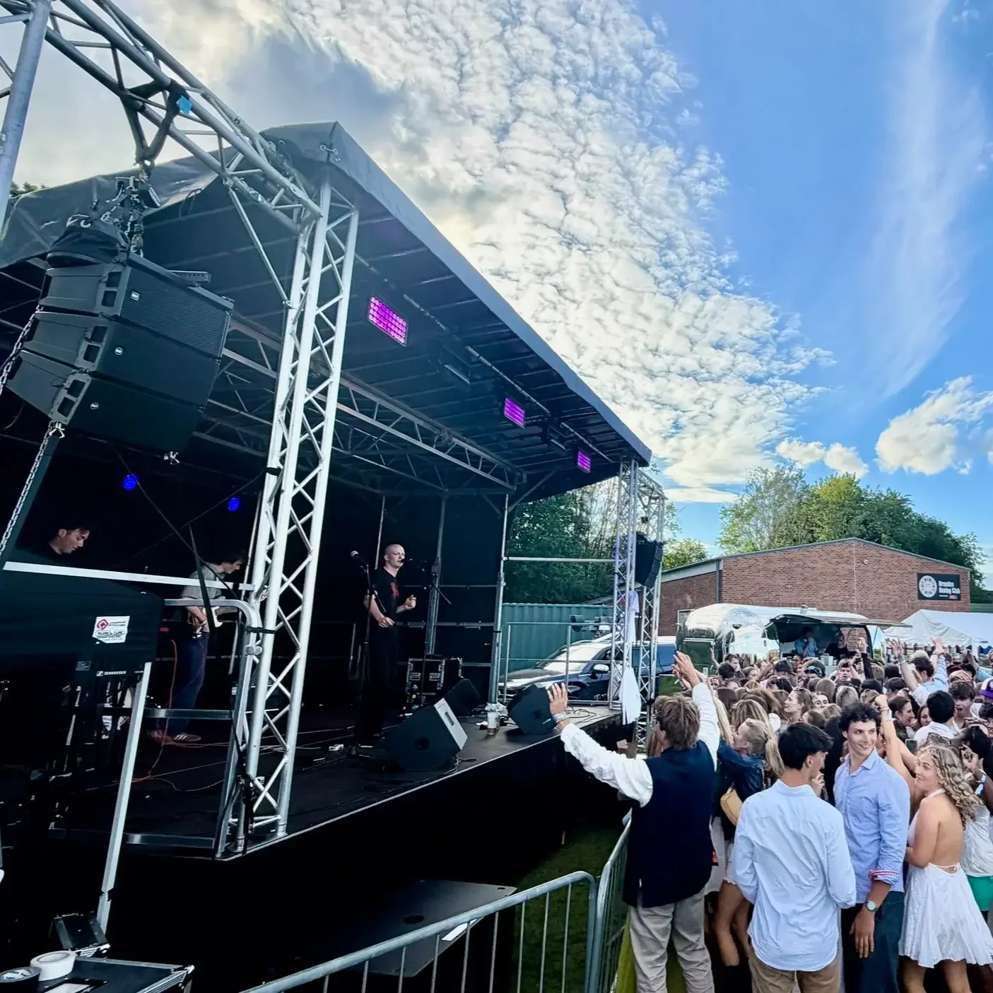 An outdoor concert with a stage, performers, and a crowd of young people dancing and enjoying the music.