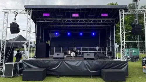 Empty outdoor concert stage with musical instruments and sound equipment, set up on a grassy area, with trees and cloudy sky in the background.